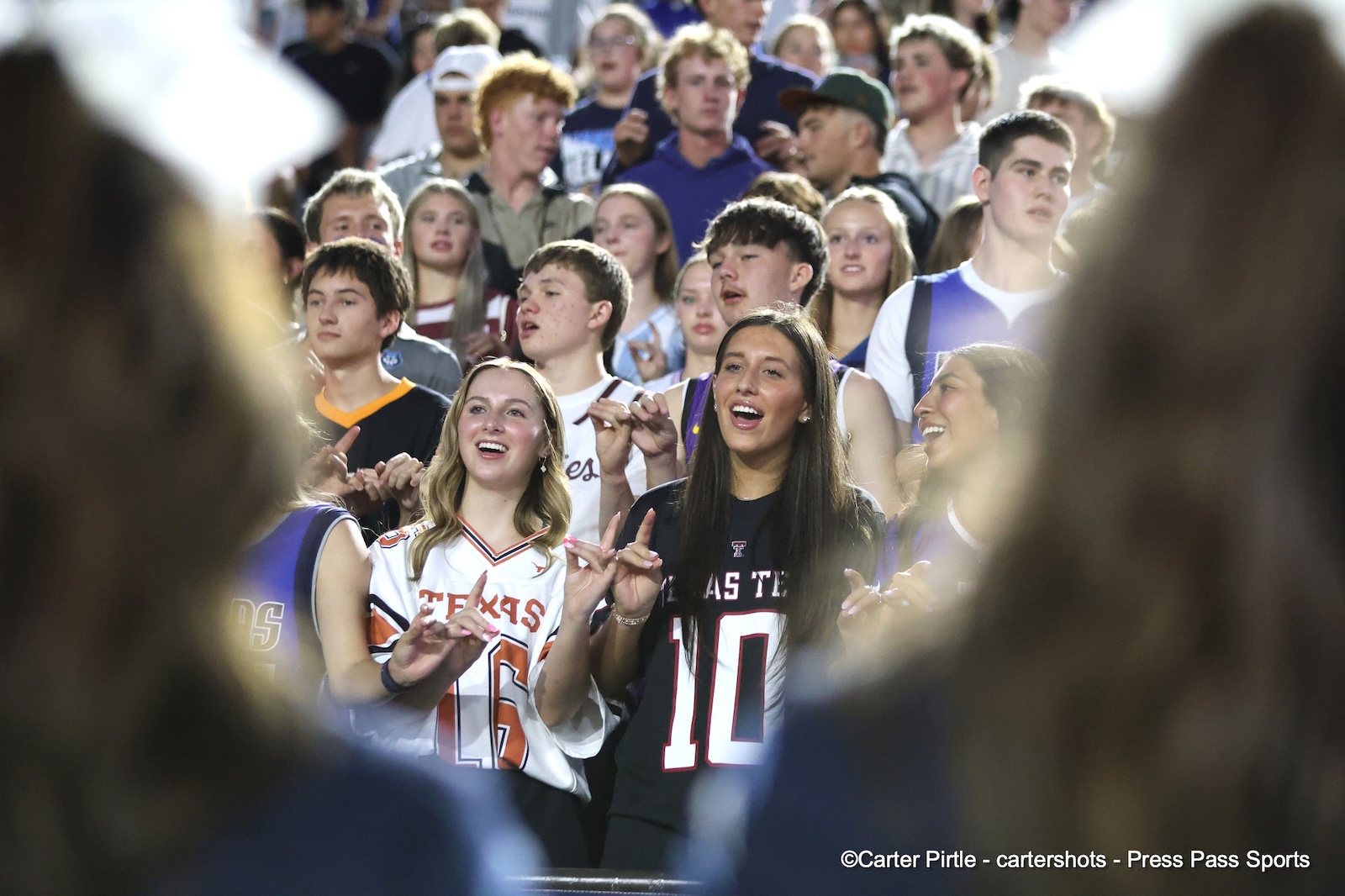 A group of smiling sports fans in jerseys cheer from the stands, with three women in the front center wearing Texas/college uniforms.
