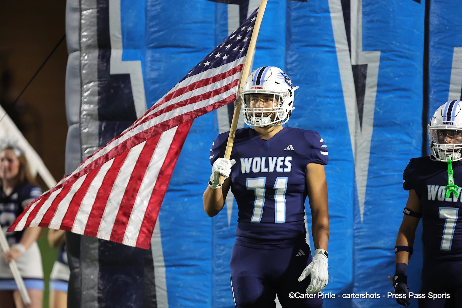 Wolves football player in navy uniform (#71) holding an American flag as he runs onto the field with blue inflatables in the background