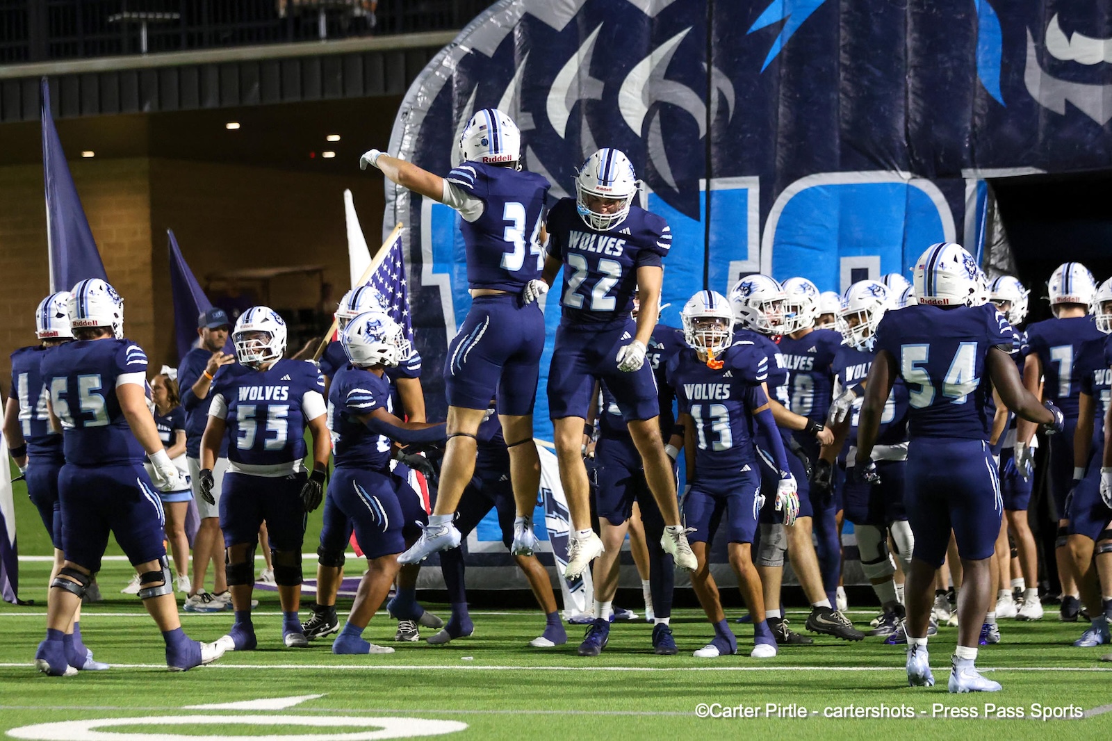 Football players in blue uniforms celebrate on a green field, two players jump midair near a large blue tunnel banner.