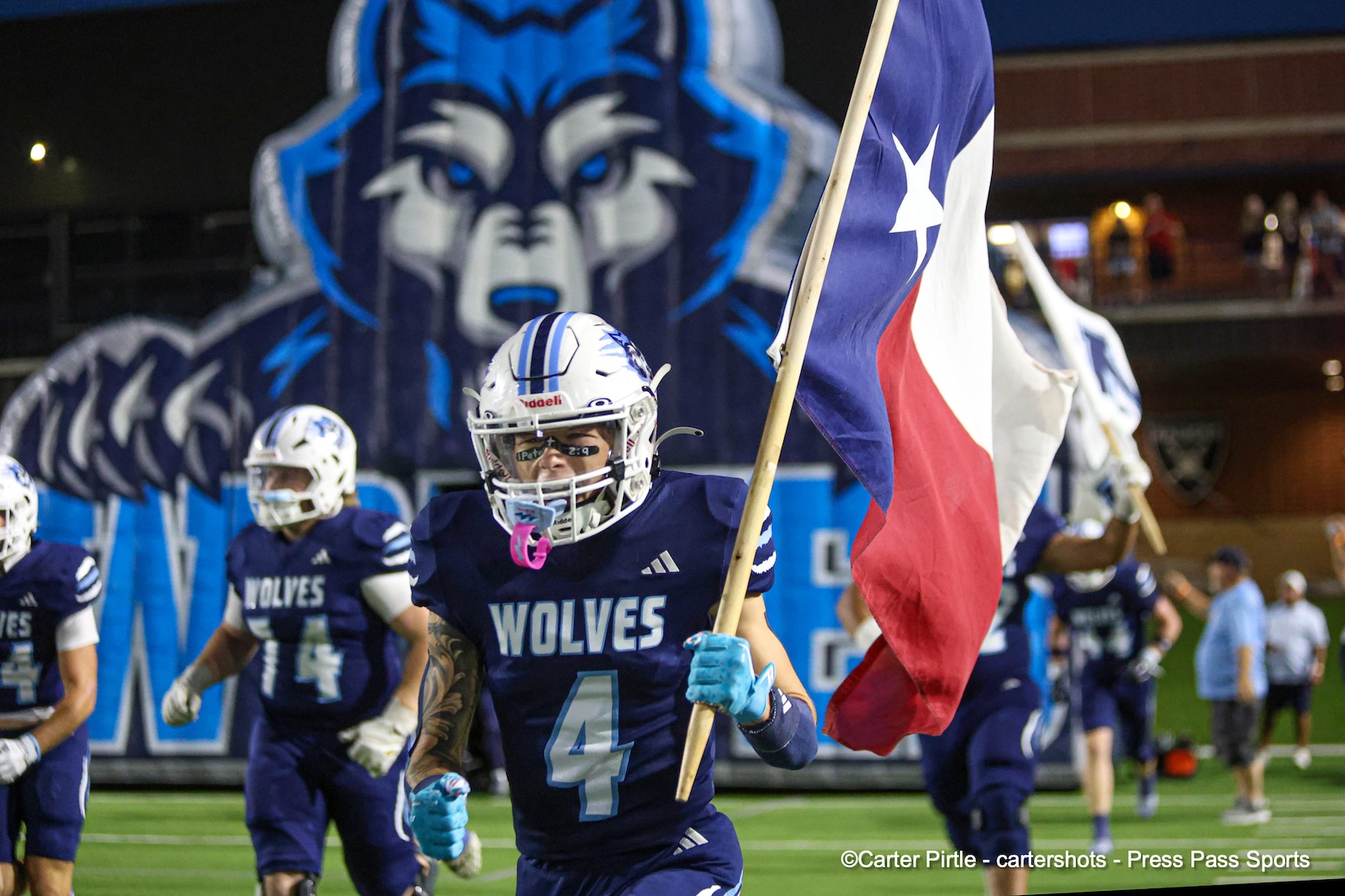 Wolves football players run onto the field; player #4 holds a large Texas flag with a blue wolf mascot in the background.