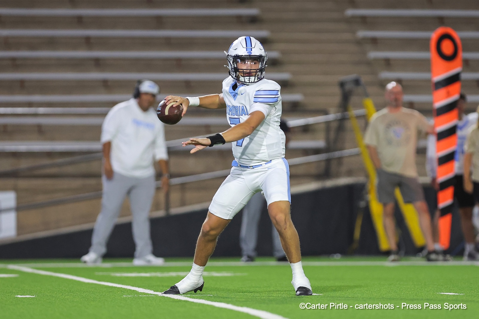 Quarterback in a white-and-blue uniform drops back to pass during a football game, gripping the ball and scanning the field.
