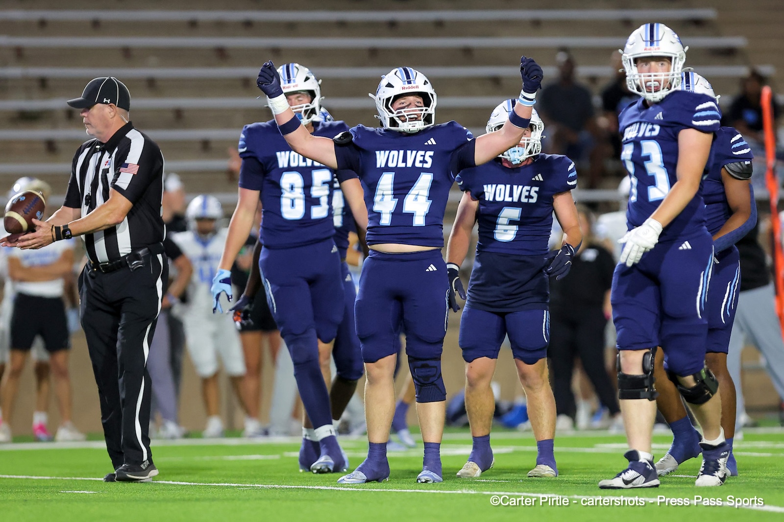 Blue-clad Wolves football players celebrate on the field with arms raised, referee to the side.