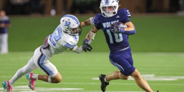 Two football players collide on a green turf field at night: white #32 reaching for the ball carrier in blue #10 holding the ball clutched to his chest