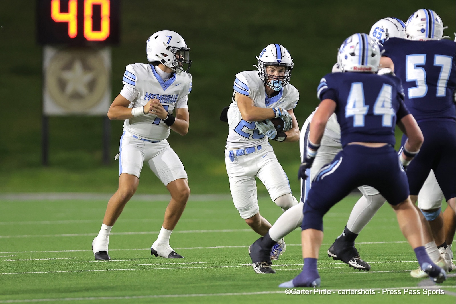 White-uniformed football players on a night game: one running with the ball while teammates block and blue-defenders close in; a 40 on the scoreboard in the background.