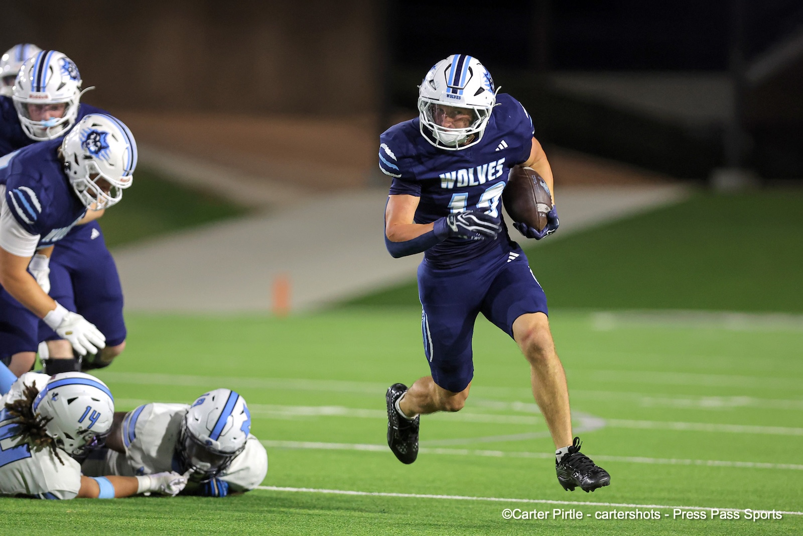 Football player in a navy Wolves uniform runs with the ball on a green field, defenders in pursuit nearby.