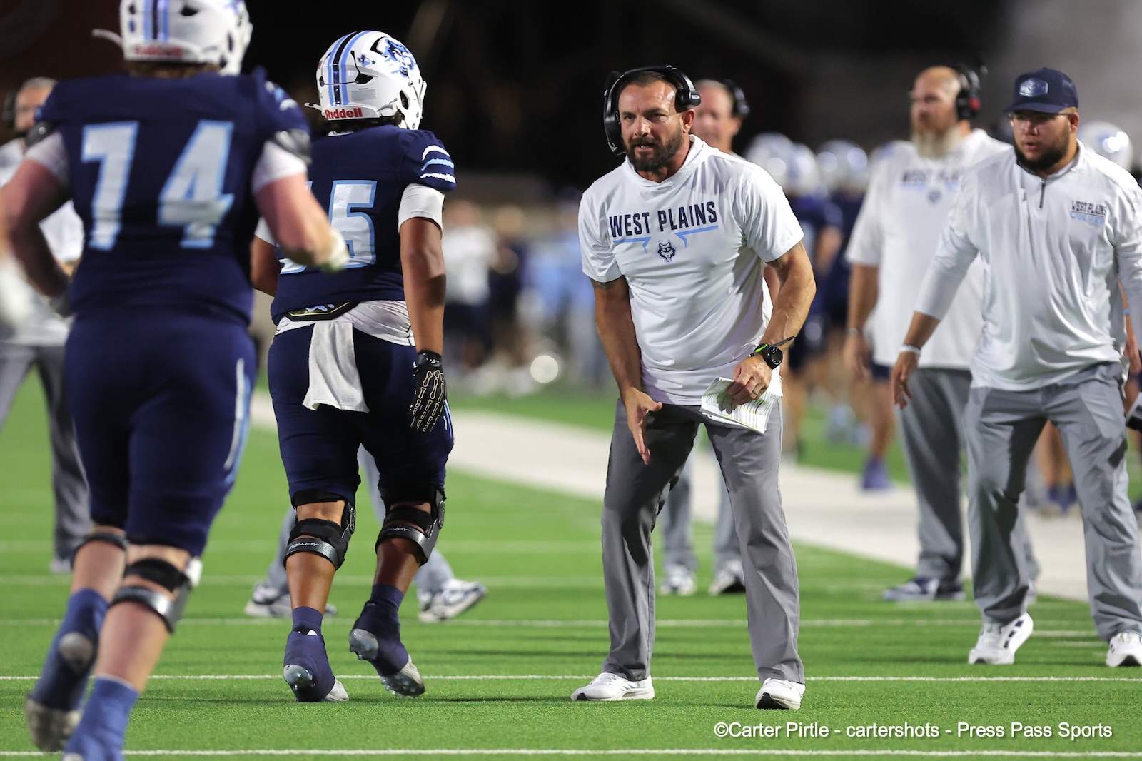 Coach wearing a headset and a'West Plains' shirt coaching players on a football field during a game-like drill on artificial turf