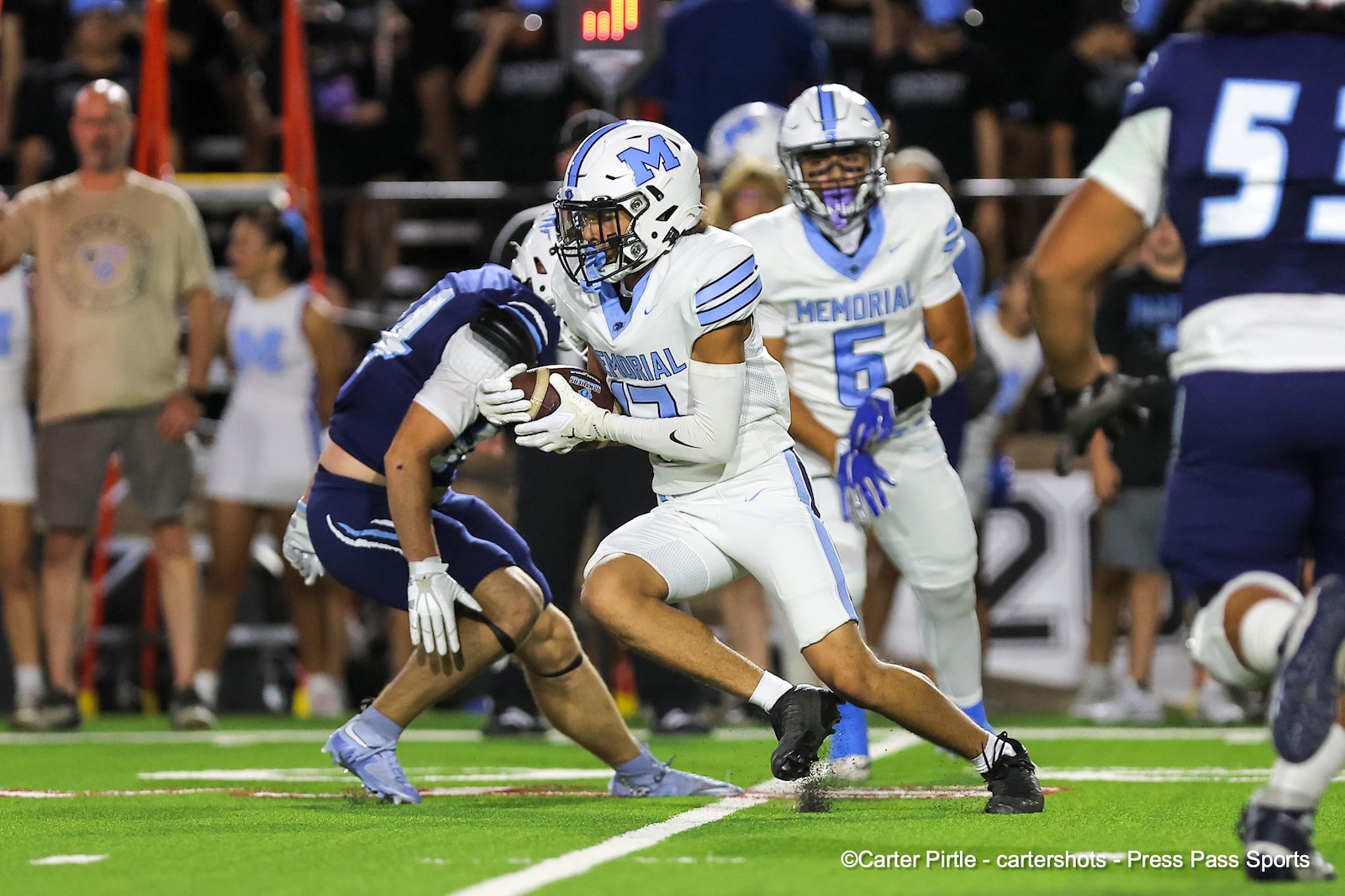 Memorial football player in white jersey with blue M runs with the ball as a blue opponent dives to tackle him on a bright turf field.