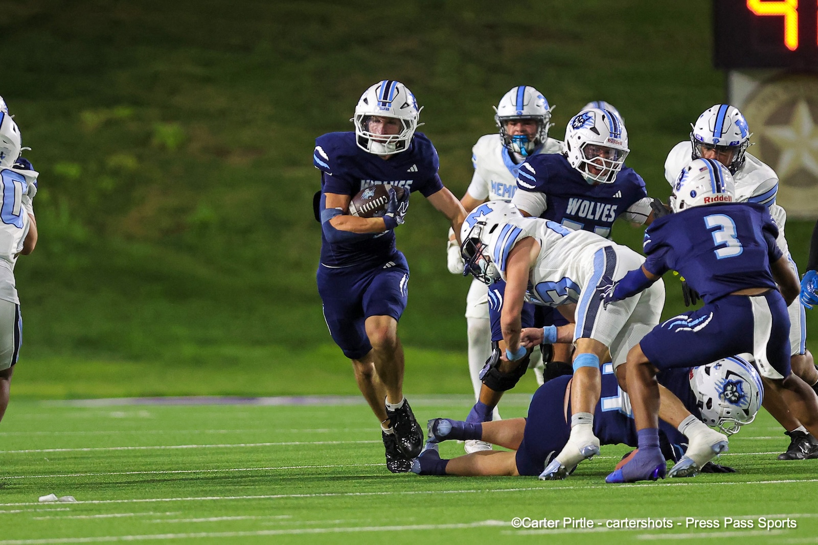 Male football player in navy uniform carries the ball while pursuing downfield, pursued by white-and-blue defenders on a green turf field behind him.