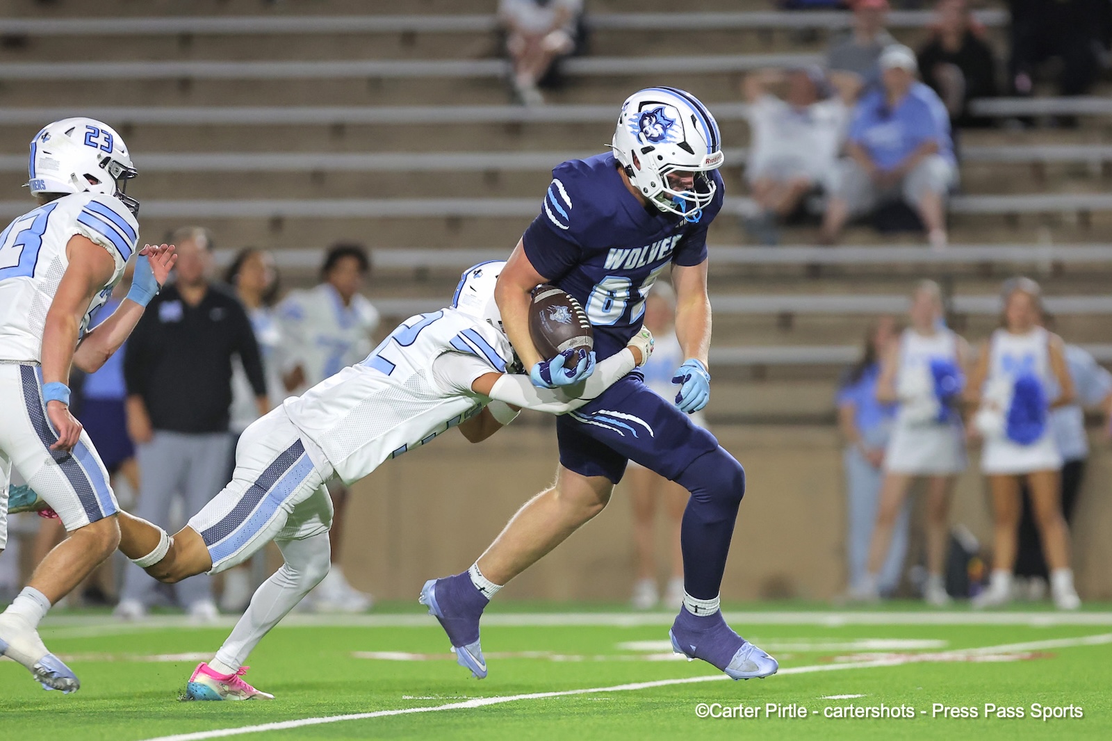 Football player in navy blue'WOLVES' jersey runs with ball as white-clad defender tackles from behind on a green turf field.