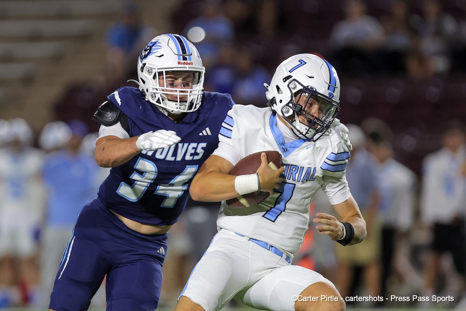 Football players in a tackle: a white-team quarterback with number 7 clutches the ball while a blue-team defender (number 54,'WOLVES') pursues him on the field.