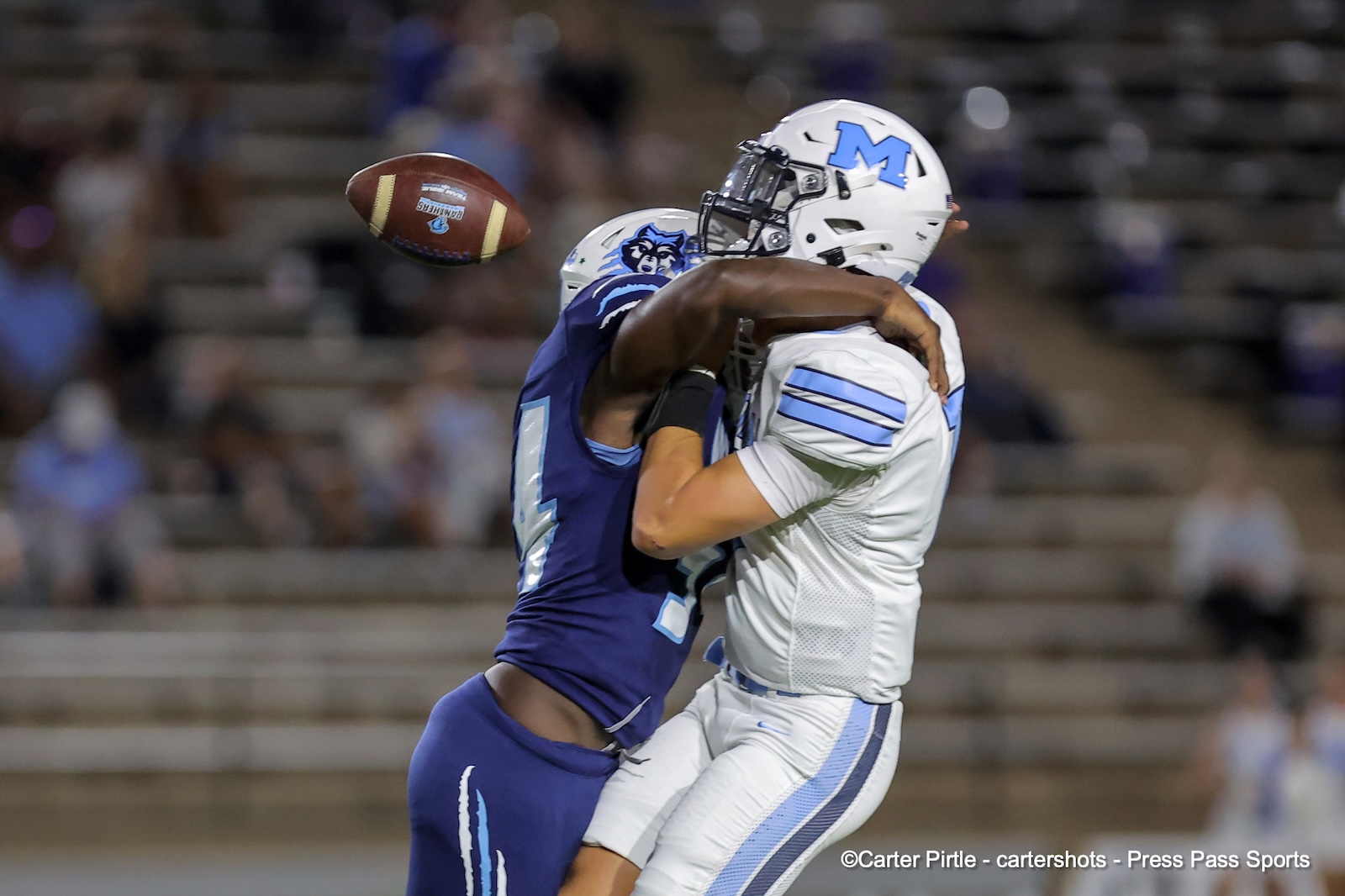 Two football players in blue and white uniforms collide for a ball in midair on a stadium field during a play.