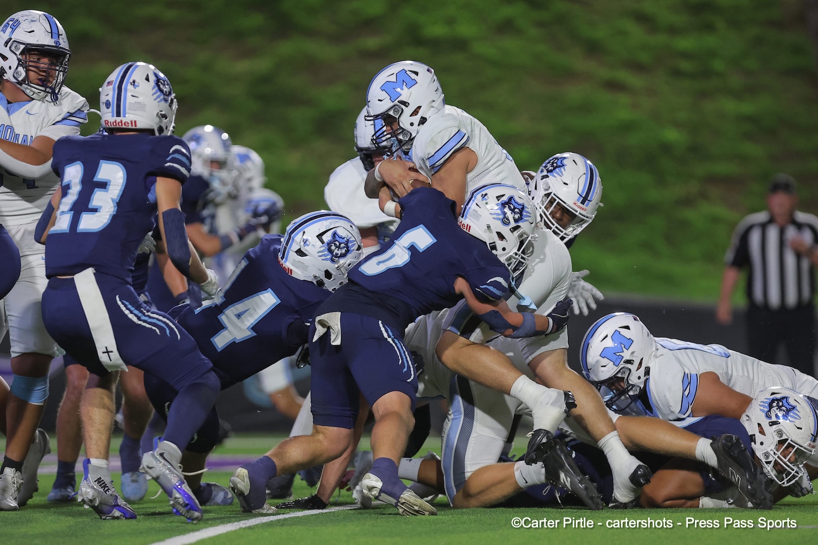 Football game action at the line of scrimmage: a ball carrier in white fights through blue-clad defenders surrounded by teammates.