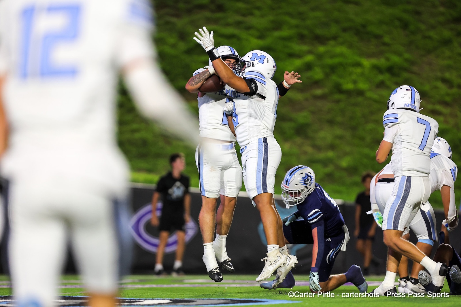 Two football players in white and light blue jerseys jump to celebrate or contest a catch on a green field as teammates rail nearby.