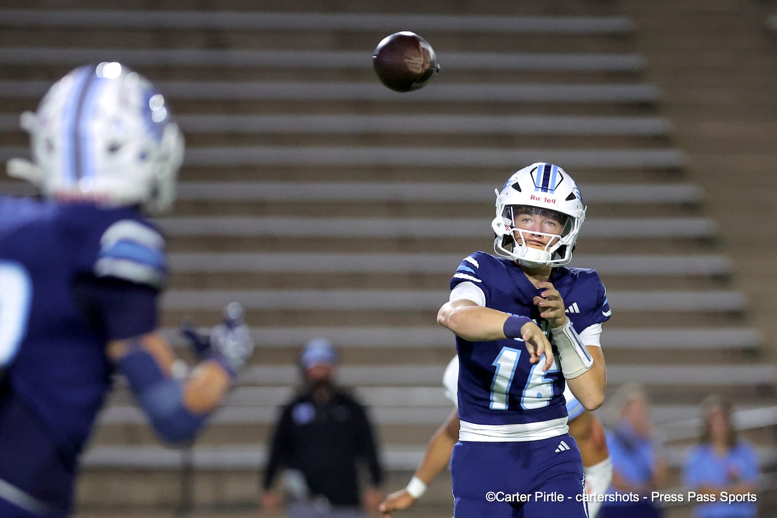 Quarterback in blue uniform mid-throw, football in air, as teammates watch on a field with bleachers in the background