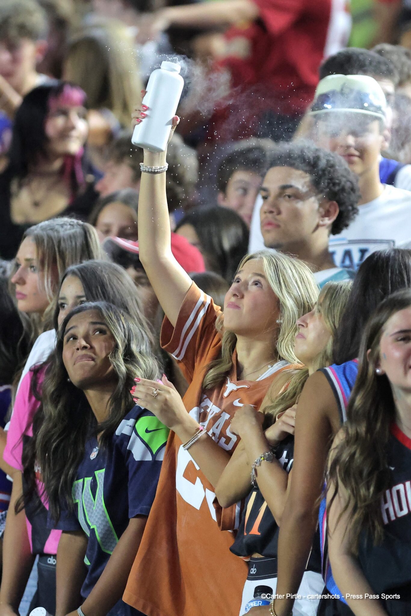 20260903-Memorial-WestPlains-FB-029 | Press Pass Sports Young woman in an orange Texas jersey raises a white spray bottle, releasing a mist into a crowded stadium crowd.