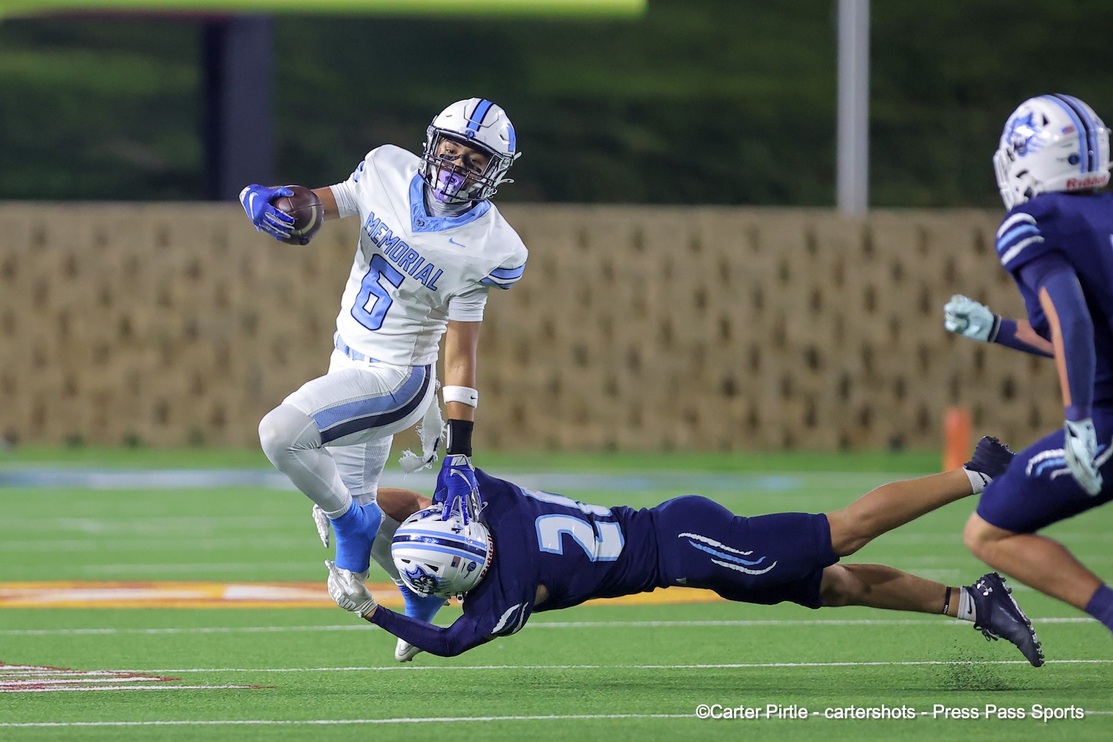 Memorial #6 football player in white jersey runs with the ball as a defender in blue dives to tackle him on a green field.
