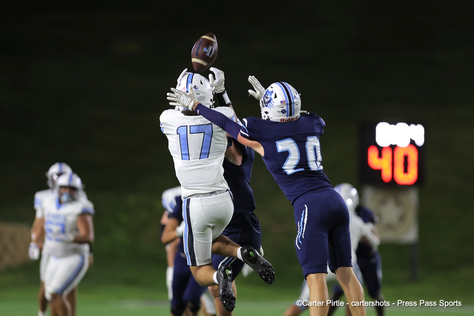 Two football players jump for a pass, white #17 reaching up as blue #20 defends nearby with a football in midair behind them