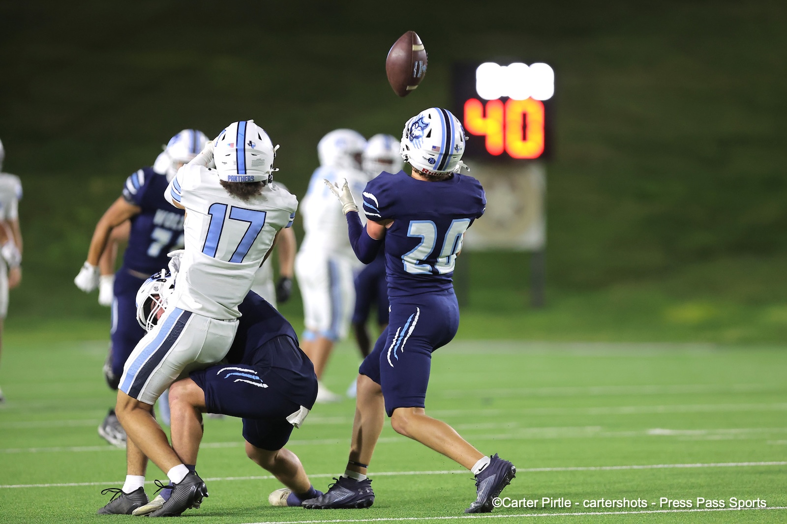 Football players in blue and white jerseys contesting a high catch as the ball sails overhead on a green turf field at night, scoreboard glowing in the background