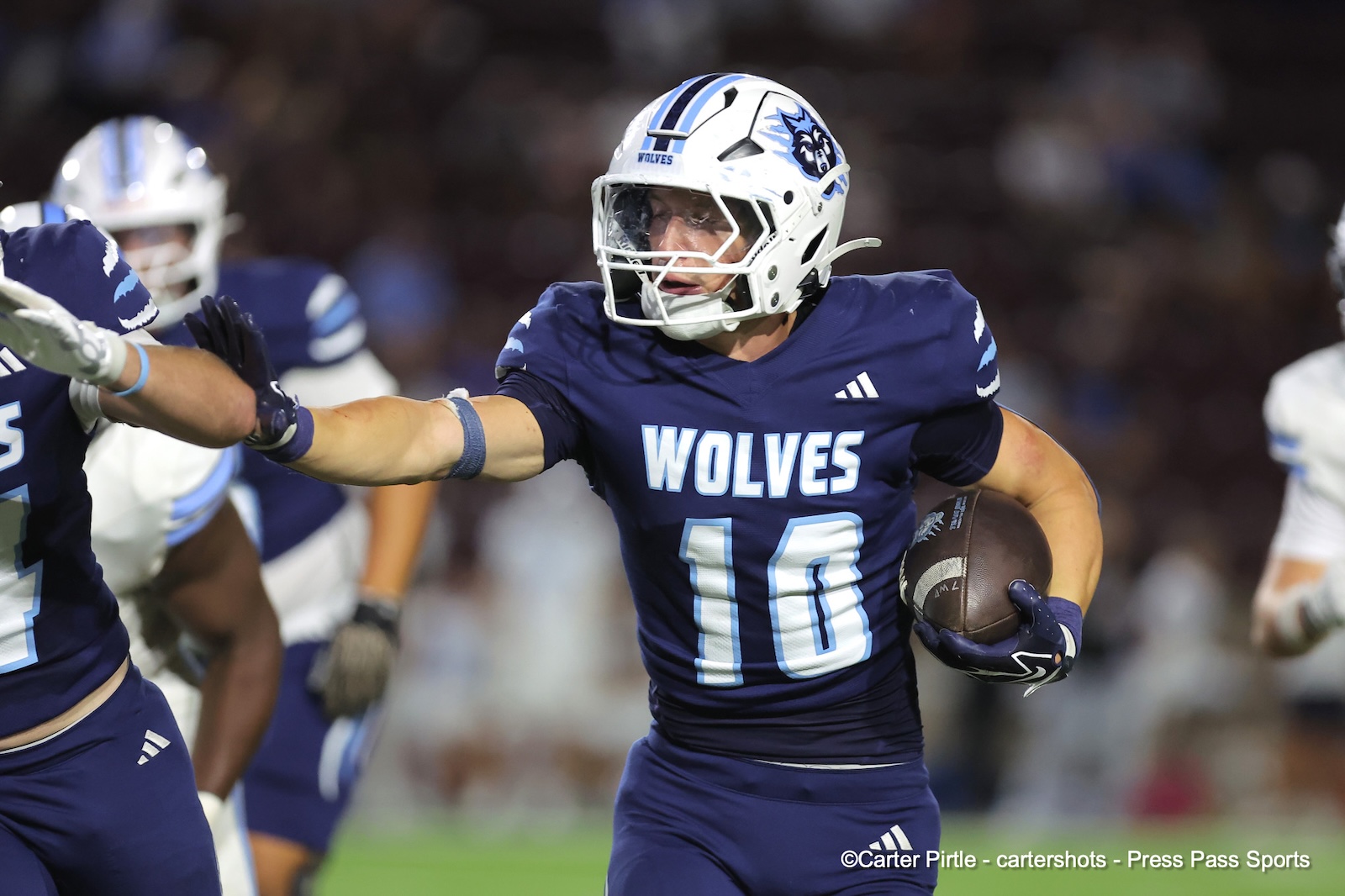 Football player in a blue Wolves uniform holds the ball and reaches forward during a game, helmet on.