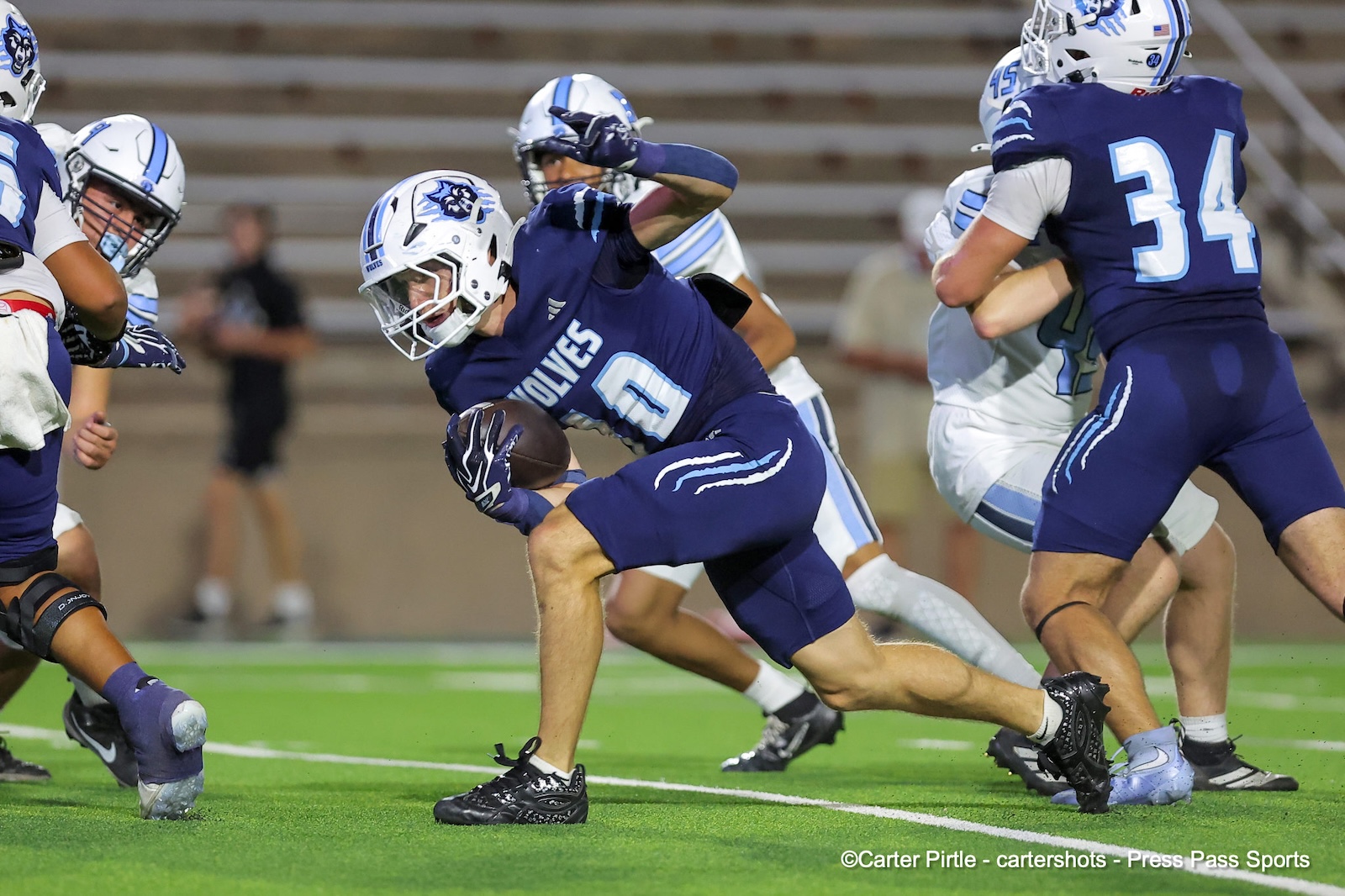 A football player in a navy blue Wolves uniform carries the ball while dodging defenders on a green turf field.