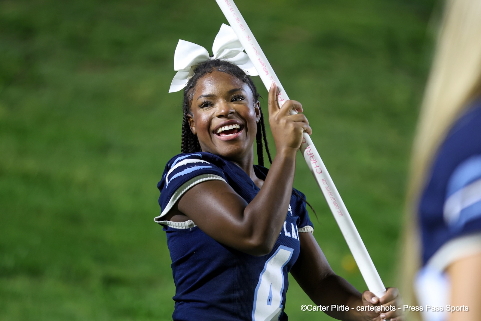 Cheerleader in a navy uniform smiles while raising a white baton on a grassy field