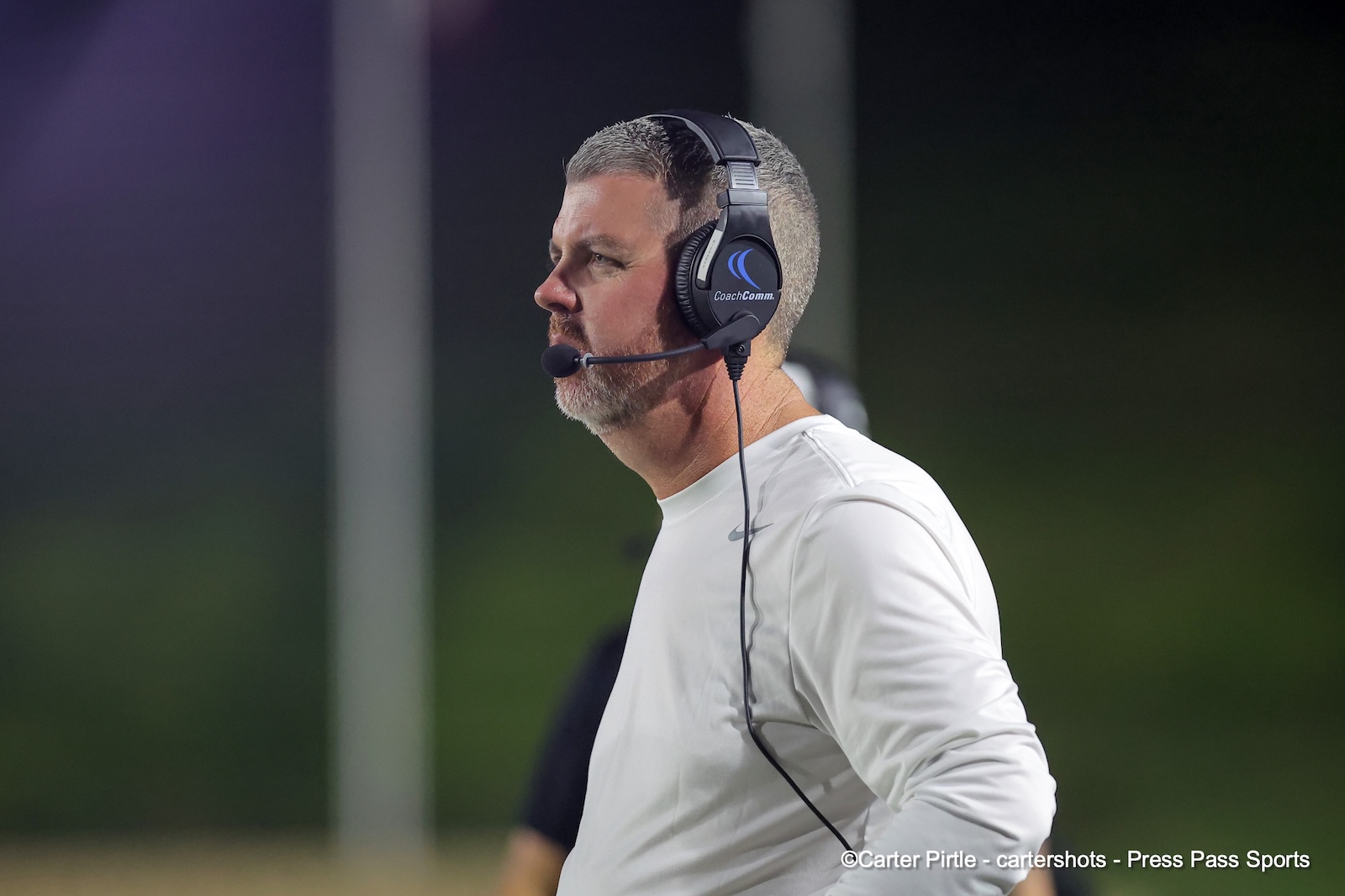 Coach wearing a black over-ear headset with microphone, standing on the sidelines in a white athletic shirt at night