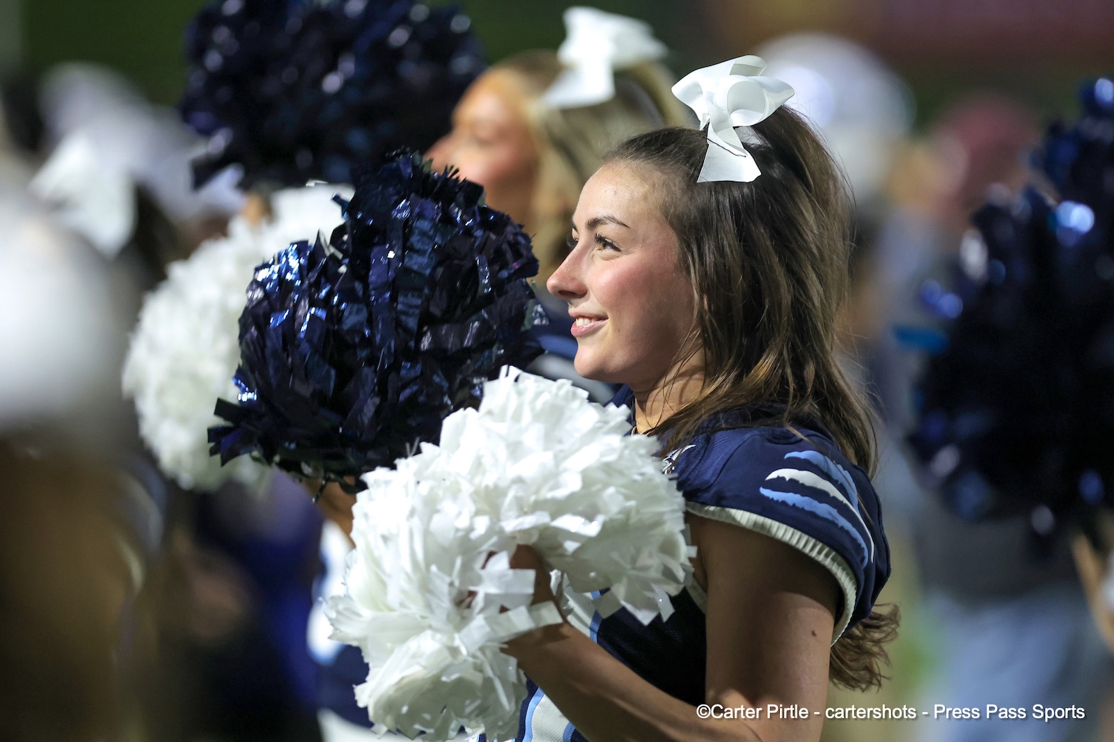 Cheerleader in a blue uniform with a white bow, smiling, holding white and navy pom-poms.