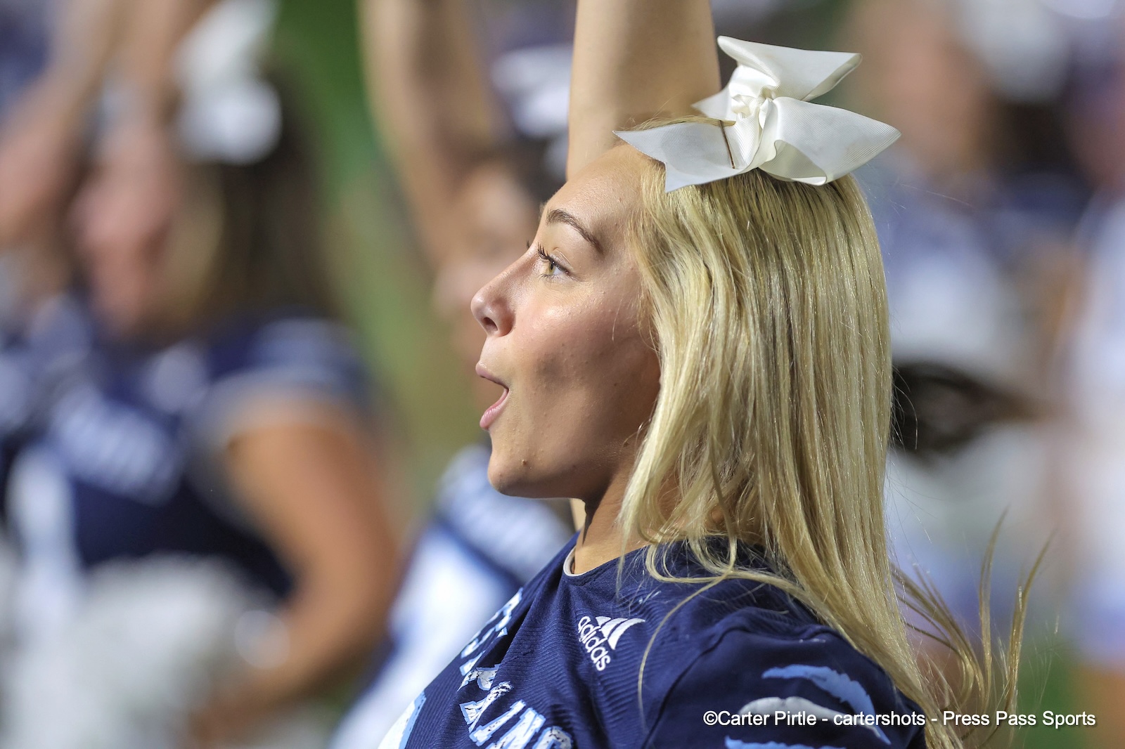 Profile view of a blonde woman cheering at a sports event, wearing a white bow and navy jersey with Adidas logo.