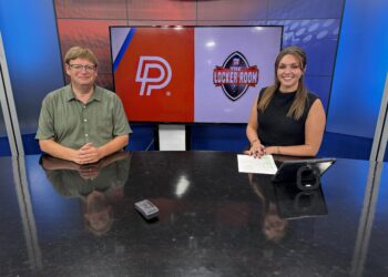 Two news anchors sit at a glass-topped desk in a studio with a large screen behind showing a DP logo and The Locker Room emblem.