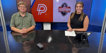 Two news anchors sit at a glass-topped desk in a studio with a large screen behind showing a DP logo and The Locker Room emblem.