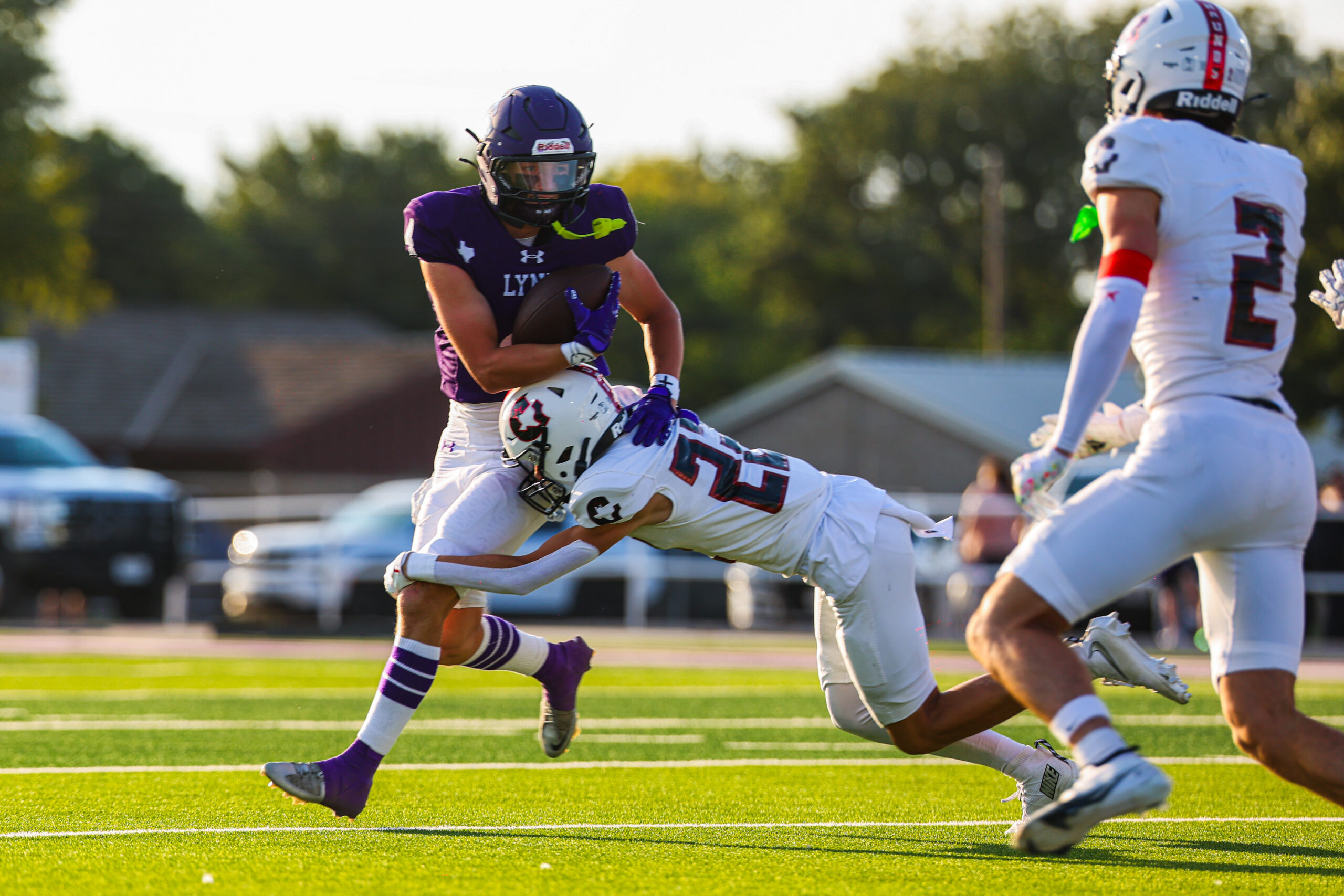 MAIRYN-02 | Press Pass Sports Purple-clad football player carrying the ball evades a defender during a daytime game on a grassy field.