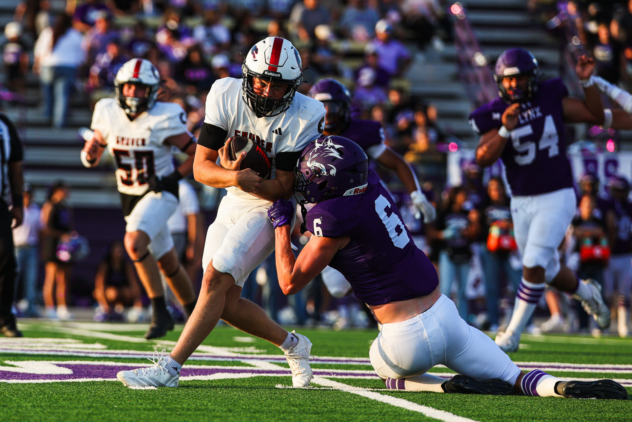 MAIRYN-06 | Press Pass Sports Football game action: a player in white shields the ball while sprinting, pursued by a purple-clad defender on a grassy field with a cheering crowd in the stands behind.