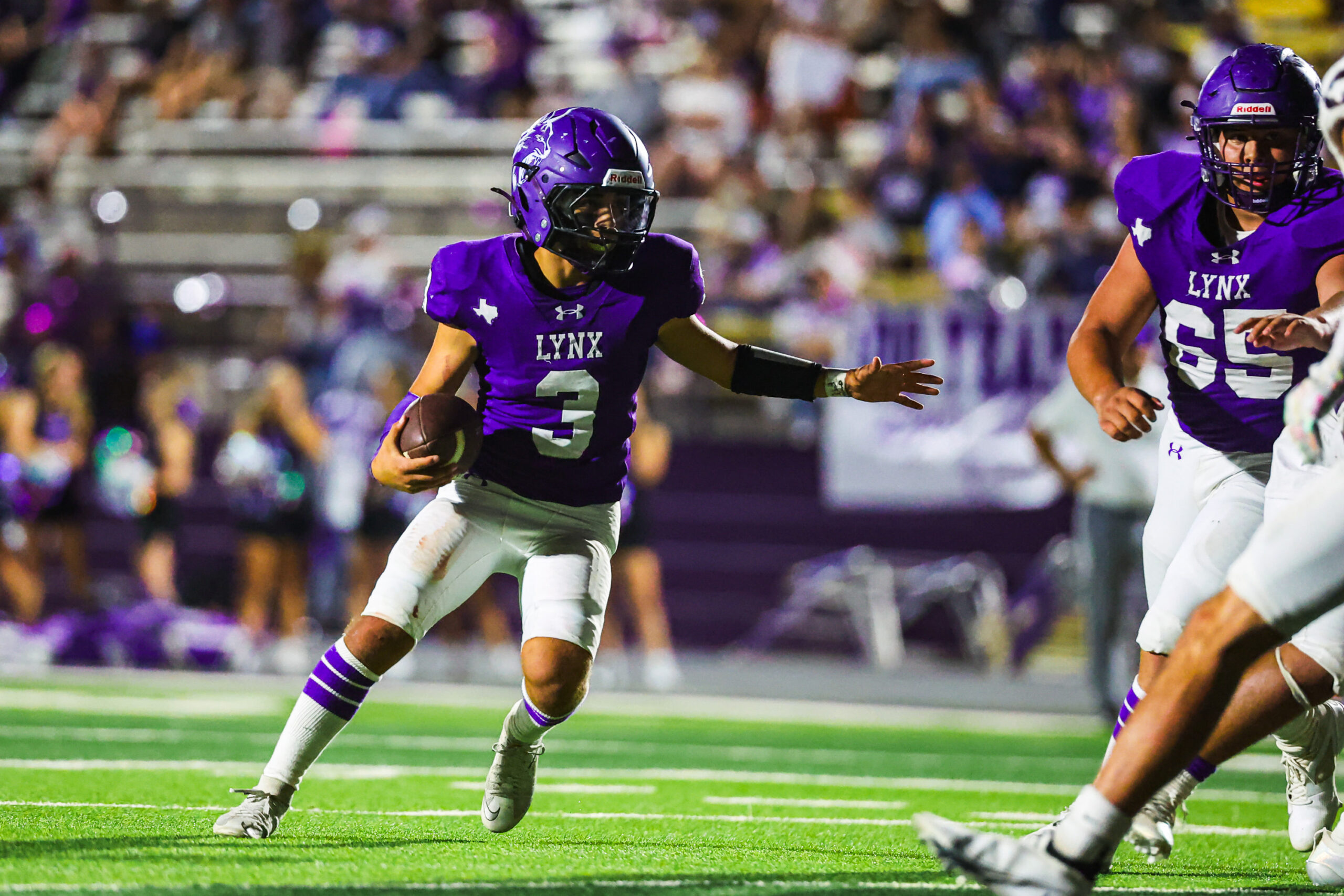 MAIRYN-15 | Press Pass Sports Two football players in purple Lynx uniforms sprint down the field; one carries the ball as a defender closes in.