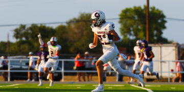 Football player in a white uniform carrying a football and sprinting down the field during a game on a green turf.