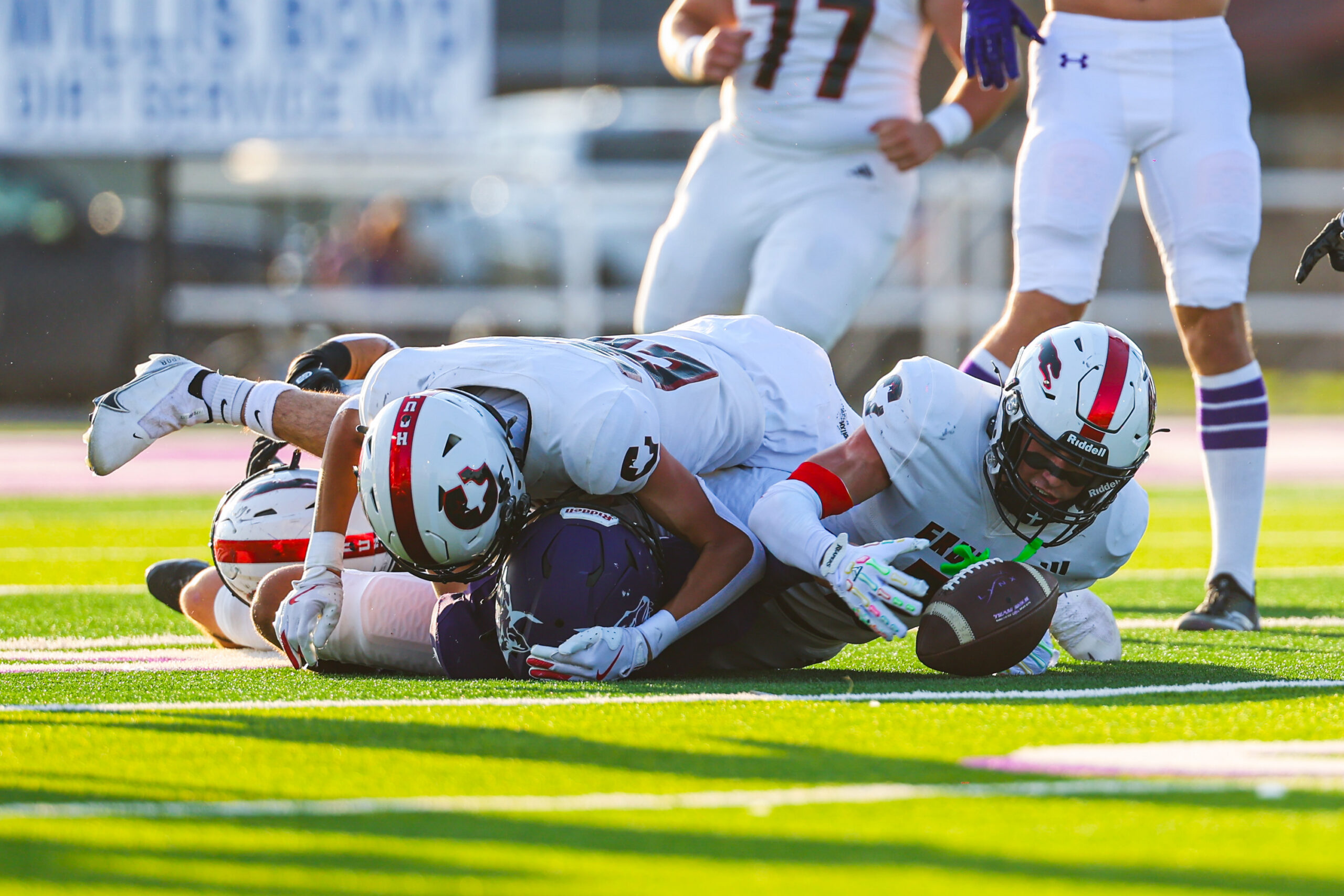 MAIRYN-20 | Press Pass Sports Football players in white uniforms pile on the grassy field during a game, helmets and padding visible as they collide for the ball momentarily downed on the turf.