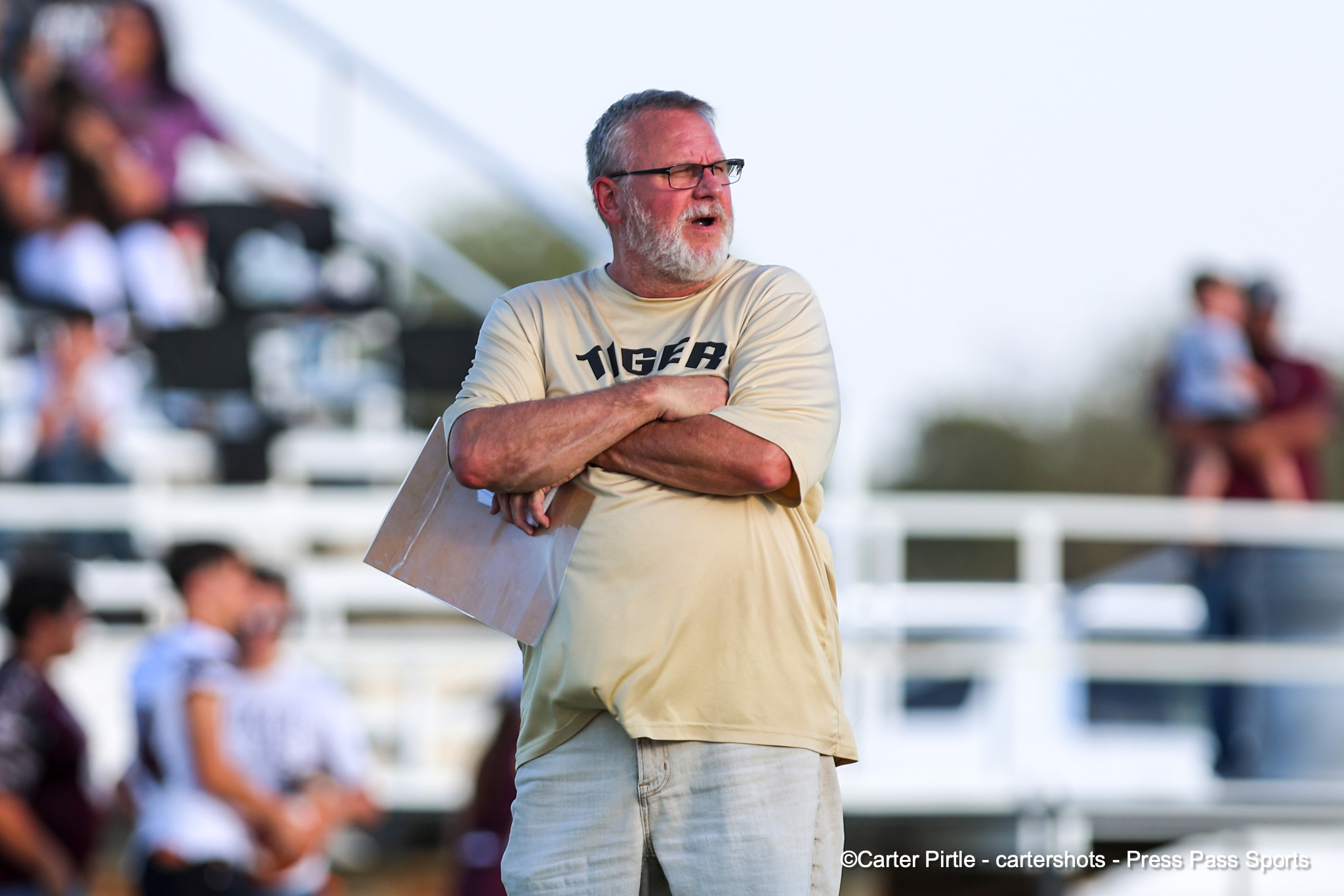 Older man with gray beard and glasses stands with arms crossed, holding a clipboard at a sports venue.