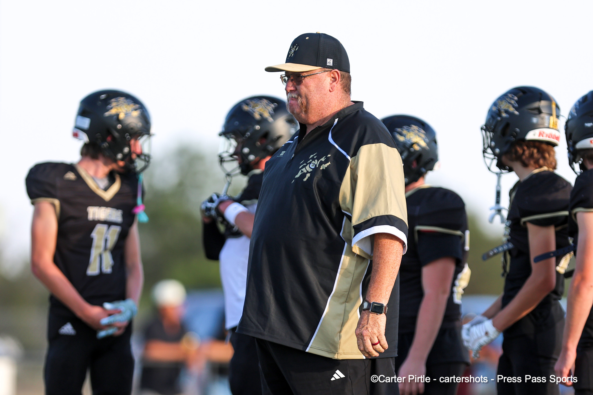 Football coach in a black and tan jacket speaks to a line of players in helmets on the field, during practice.