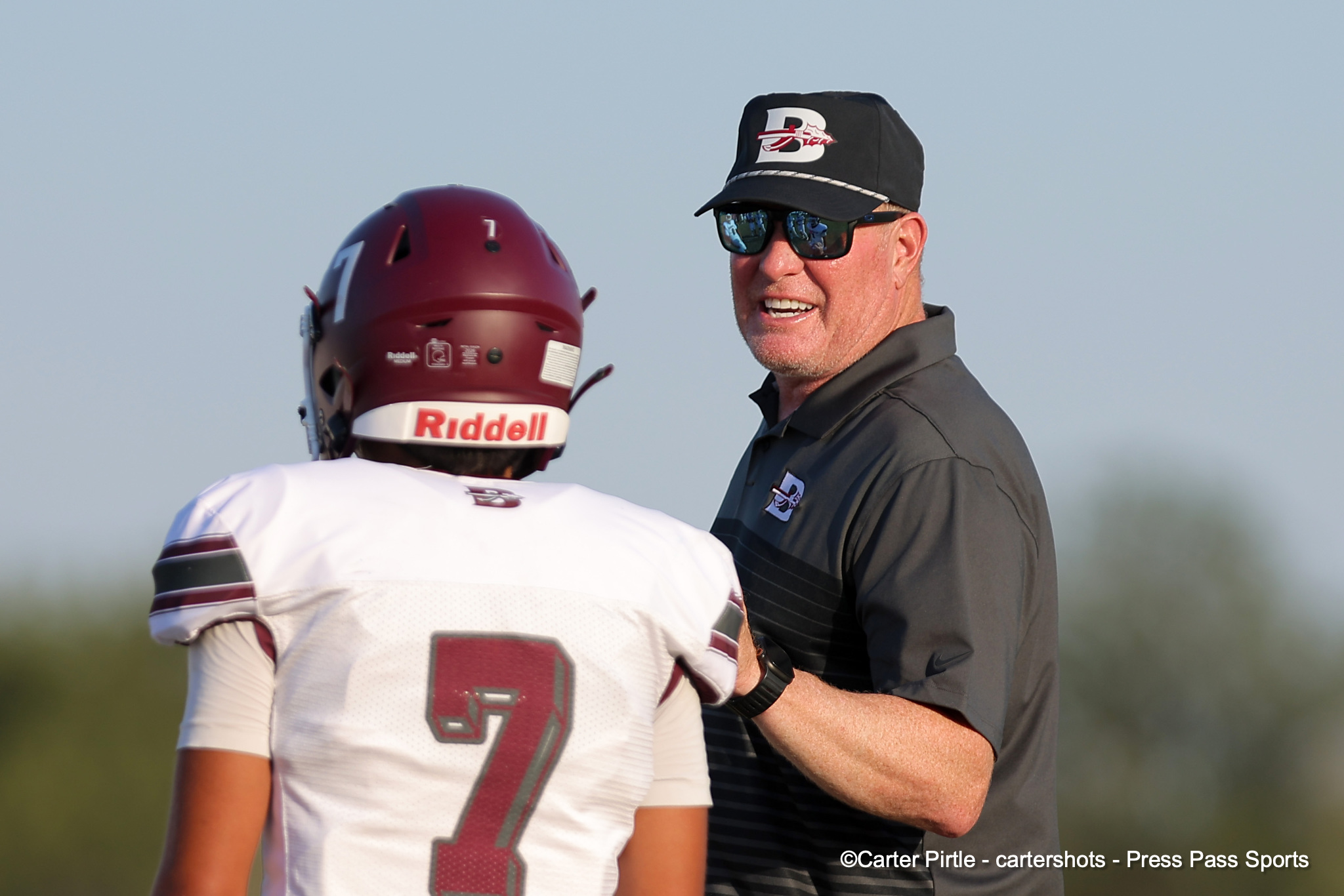 Football coach in a black cap and sunglasses speaks to a football player wearing a white jersey and a maroon helmet outdoors.
