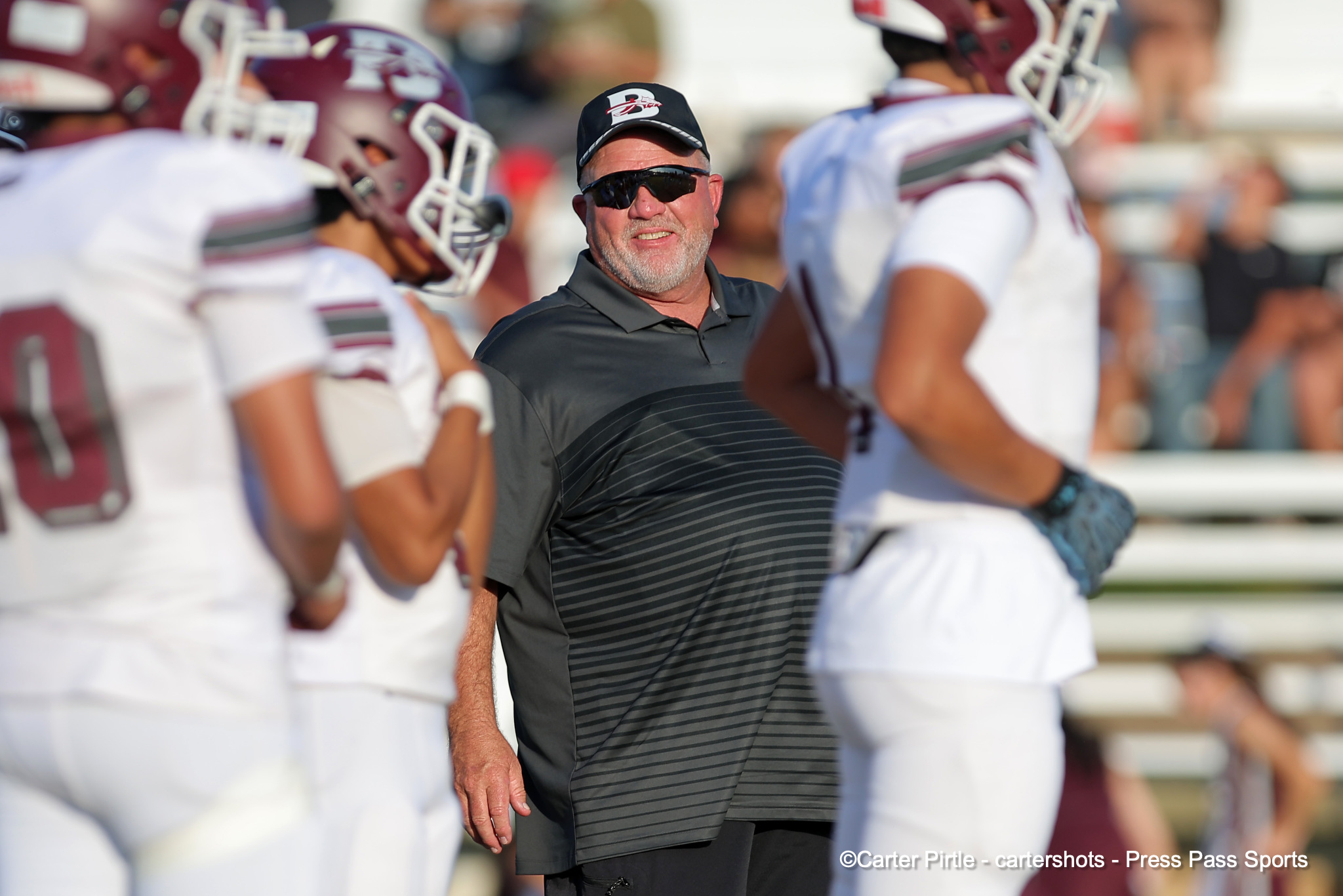 Coach wearing sunglasses and a black cap stands among football players in white uniforms on the field.