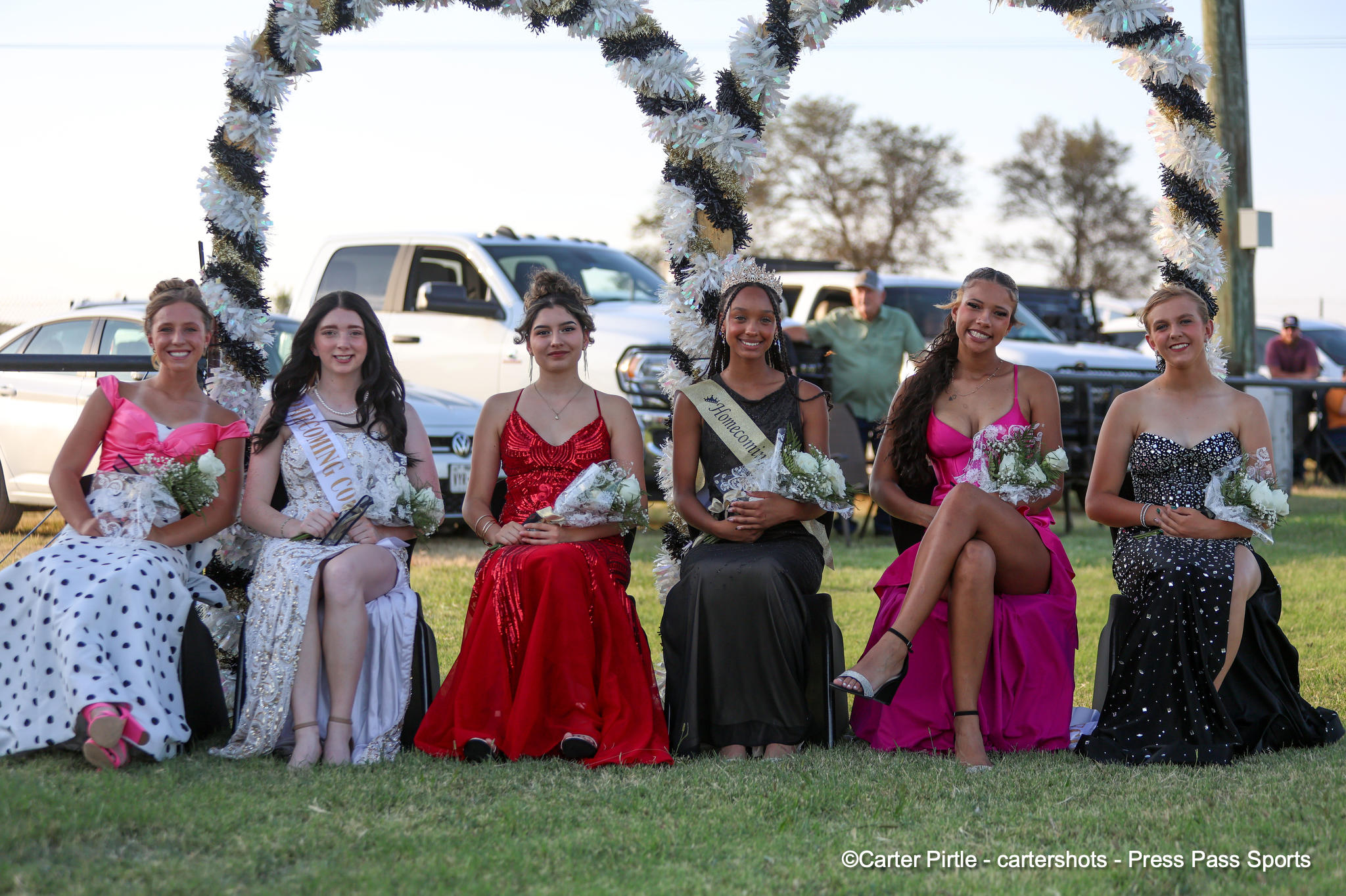 Six young women in formal dresses sit on a bench under a festive garland arch, smiling outdoors.