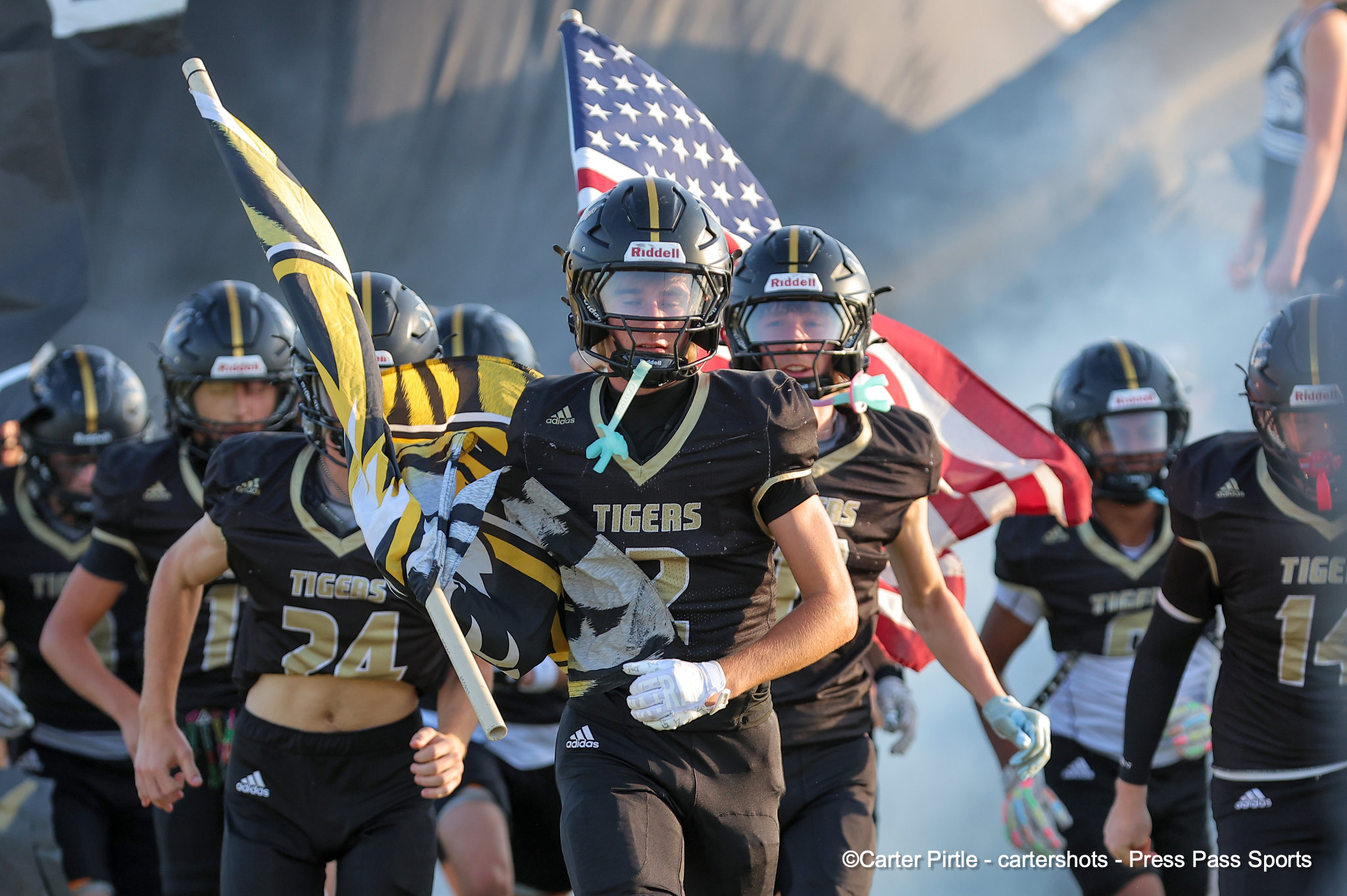 Team of Tigers football players in black uniforms sprint onto the field, an American flag visible behind them.