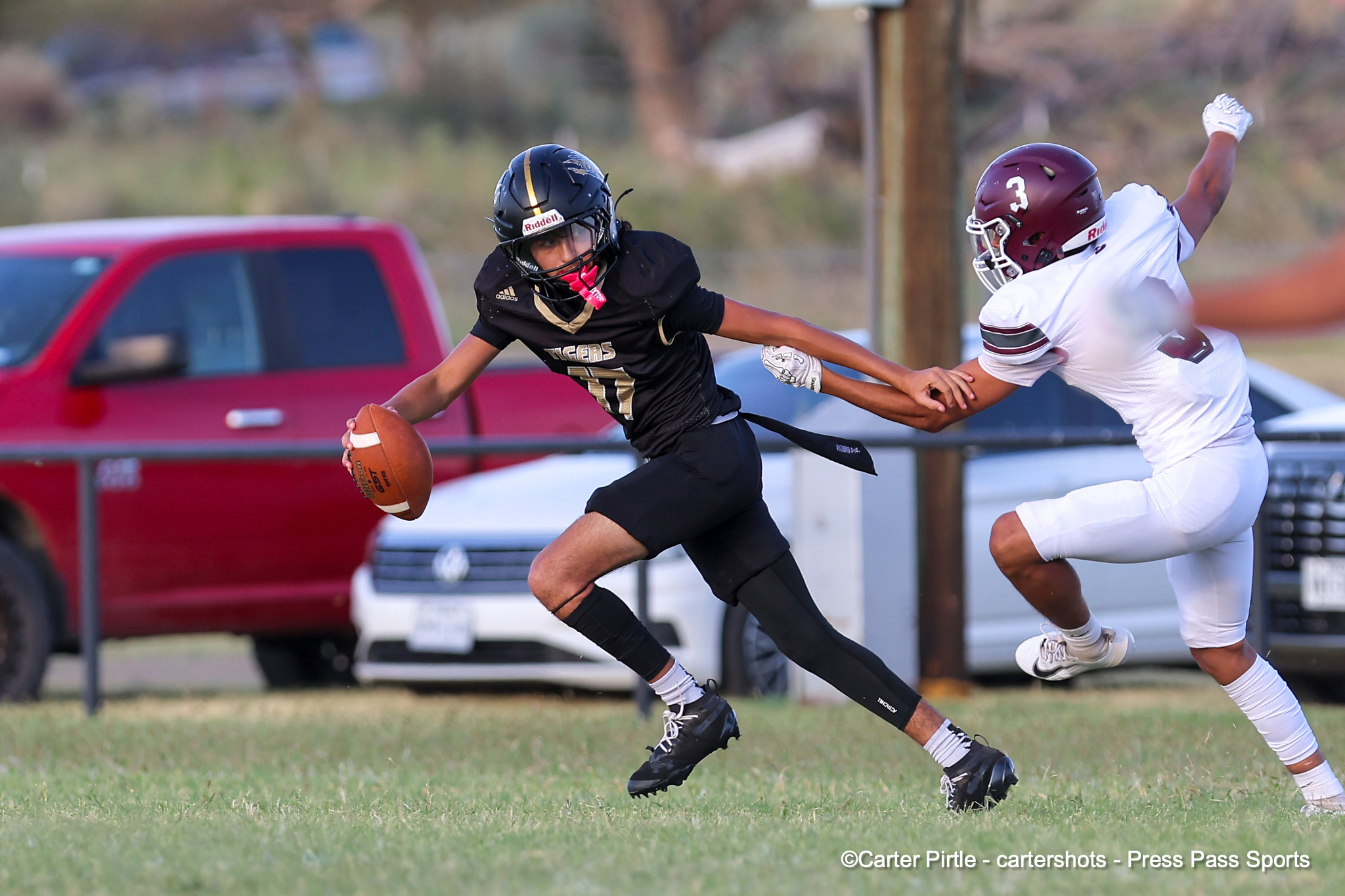 Two football players sprinting on a grassy field, one in black with the ball, being chased by a white-uniform defender. Cars and a fence visible in the background.