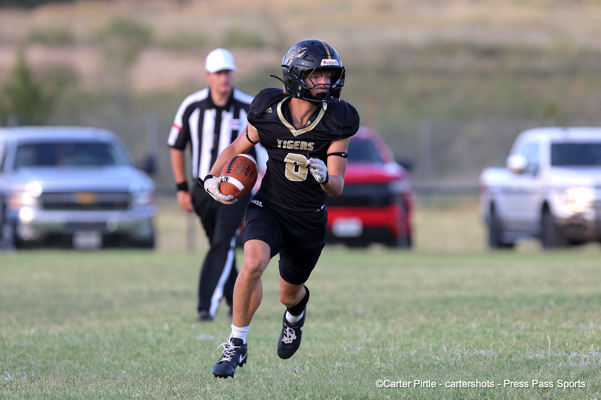 Football player in a black Tigers jersey runs with the ball on a grassy field, a referee and cars blurred in the background, during a game.