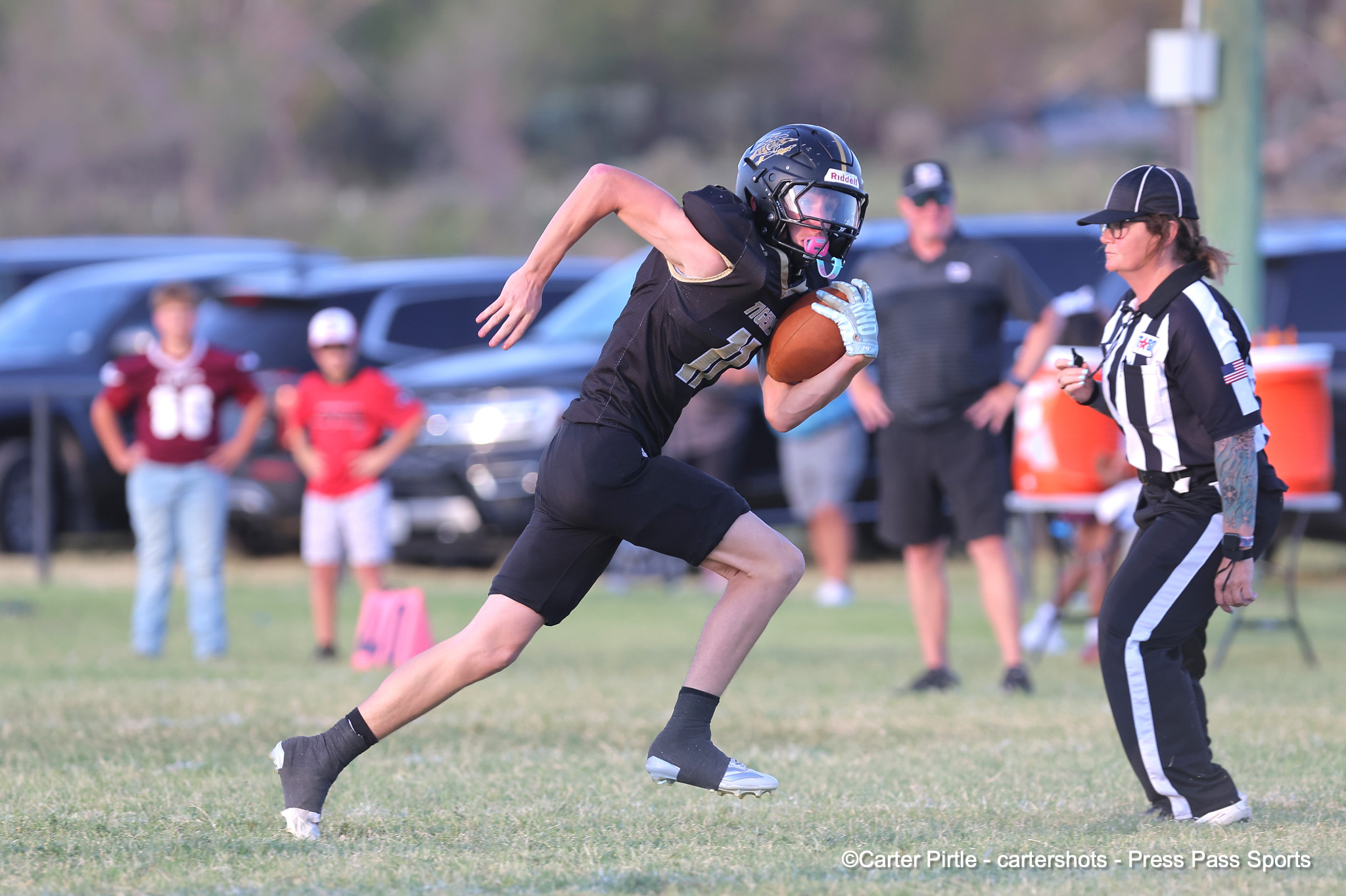 Football player in black uniform runs with ball toward end zone, referee nearby on grassy field.