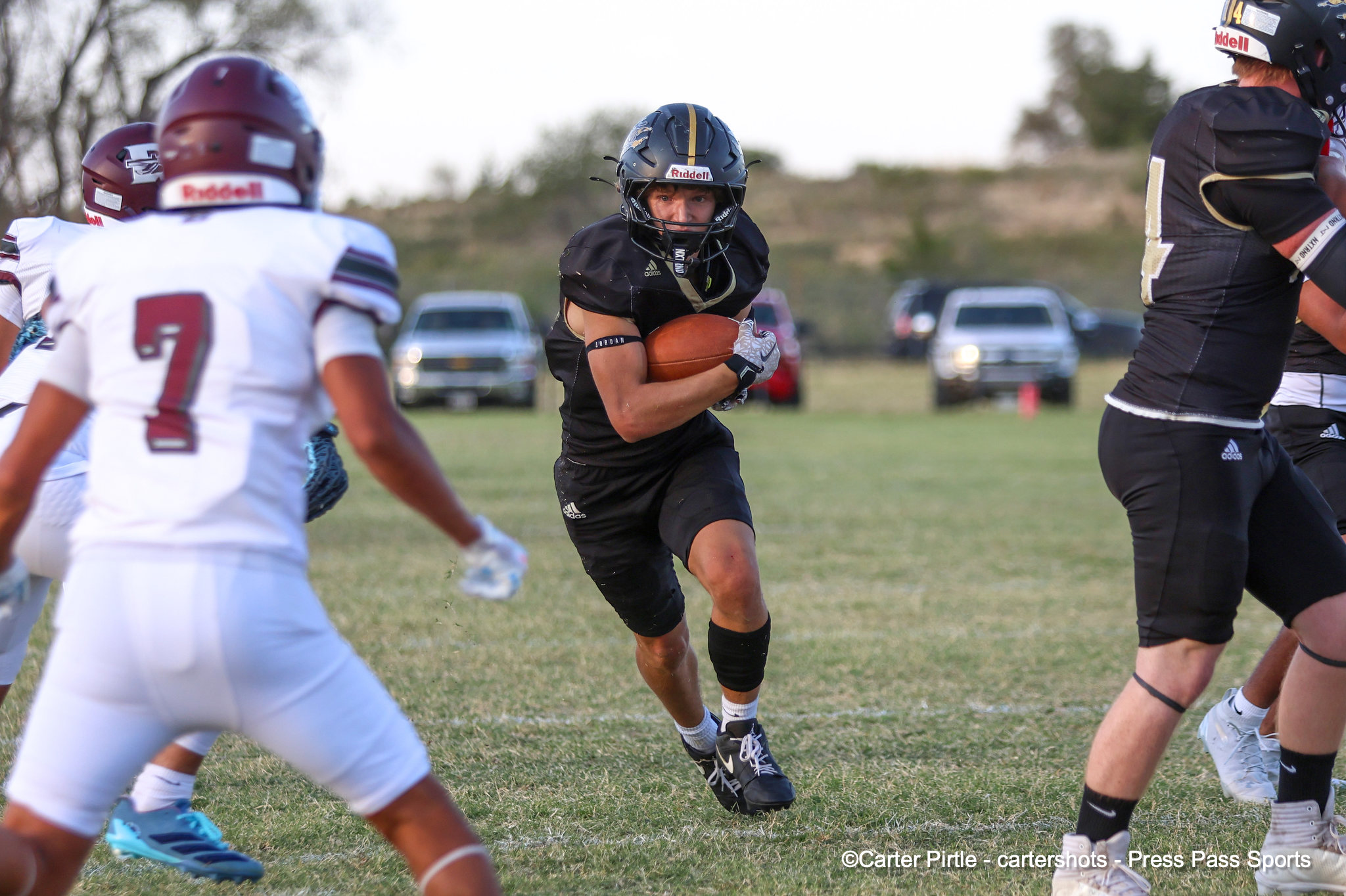 Football player in black uniform carrying the ball runs toward the camera while defenders close in on a grassy field.
