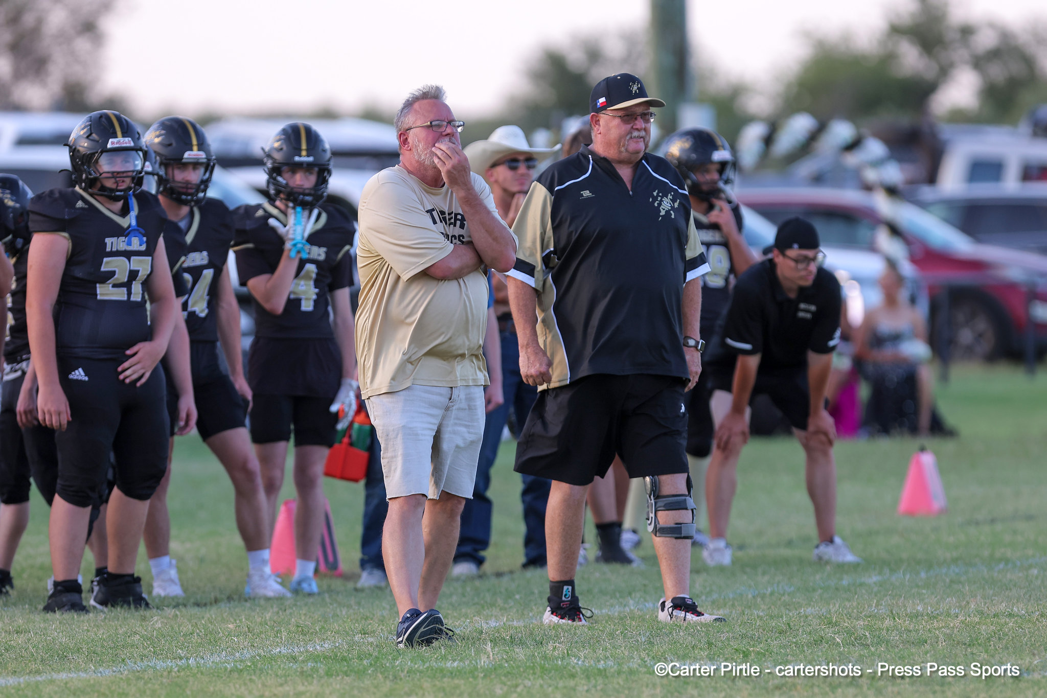Youth football team and coaches stand on a grassy field, players in black uniforms and helmets listening as two adults in front give guidance.