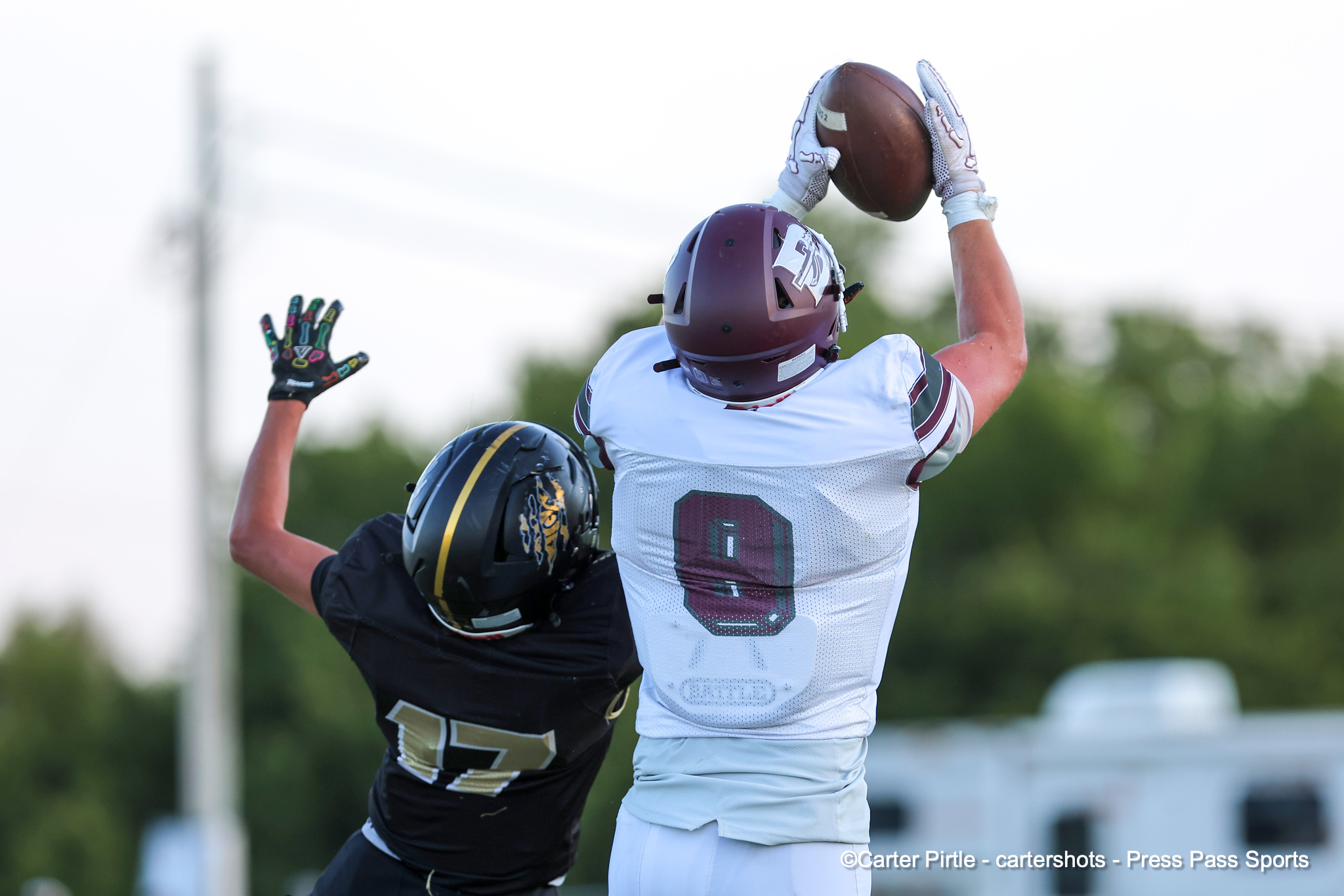 Two football players leap to catch a ball mid-air during a game; one in white uniform and maroon helmet, the other in black.