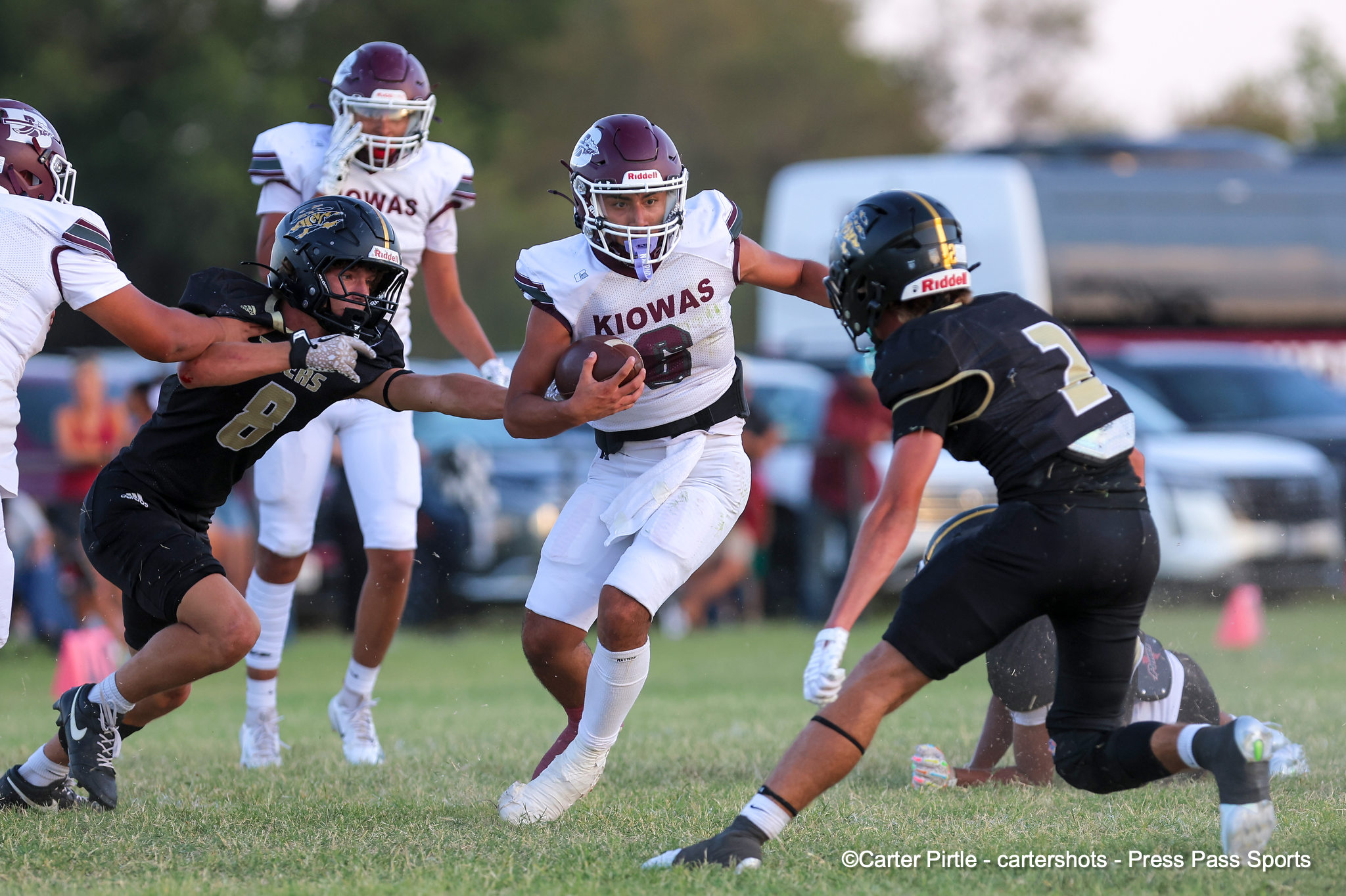 Football players in white Kiowas jerseys chase the ball on a grassy field; central runner holds the ball.