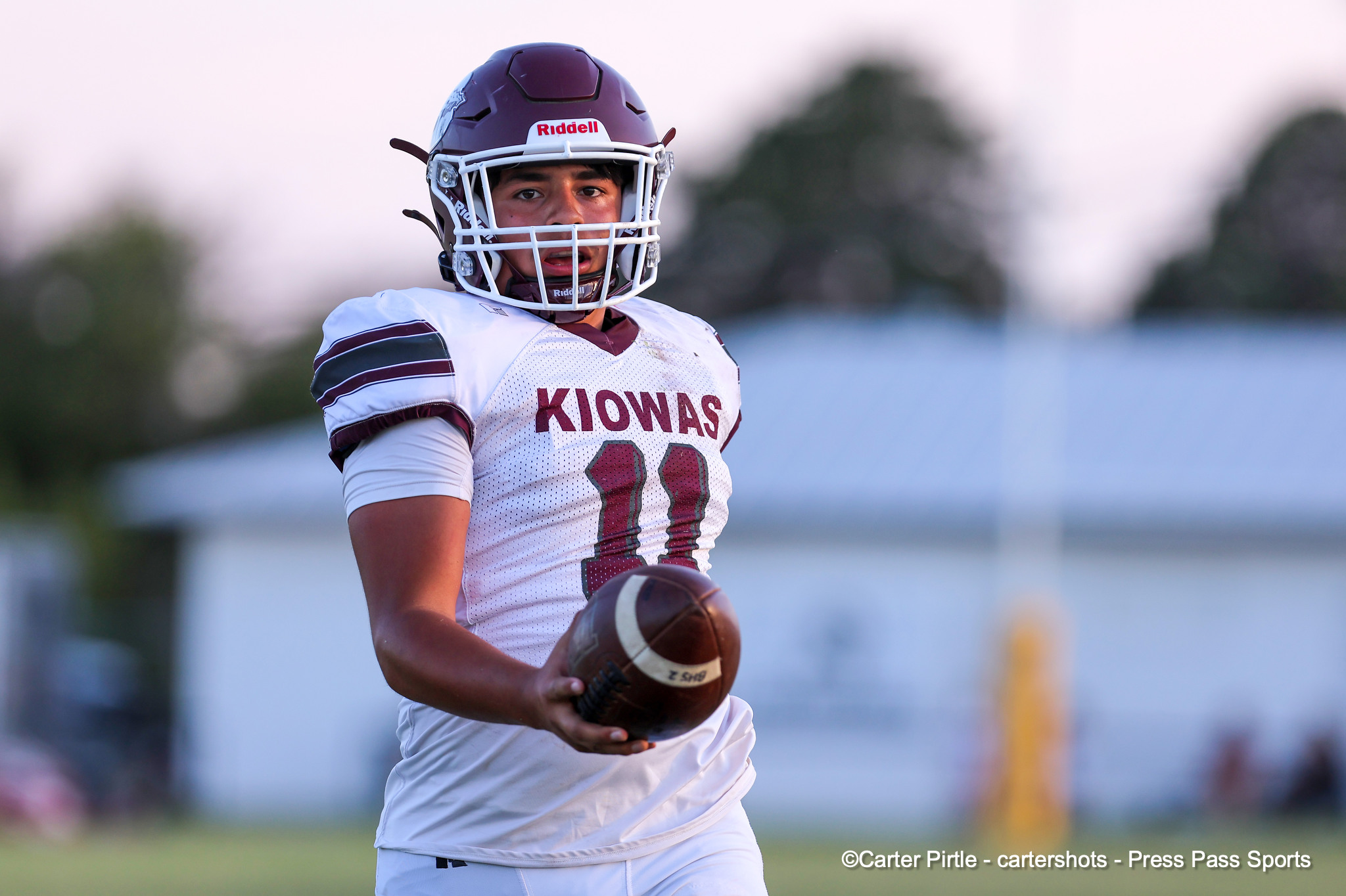 Young football player in a white and maroon Kiowas uniform holding a football on a grassy field as he runs.