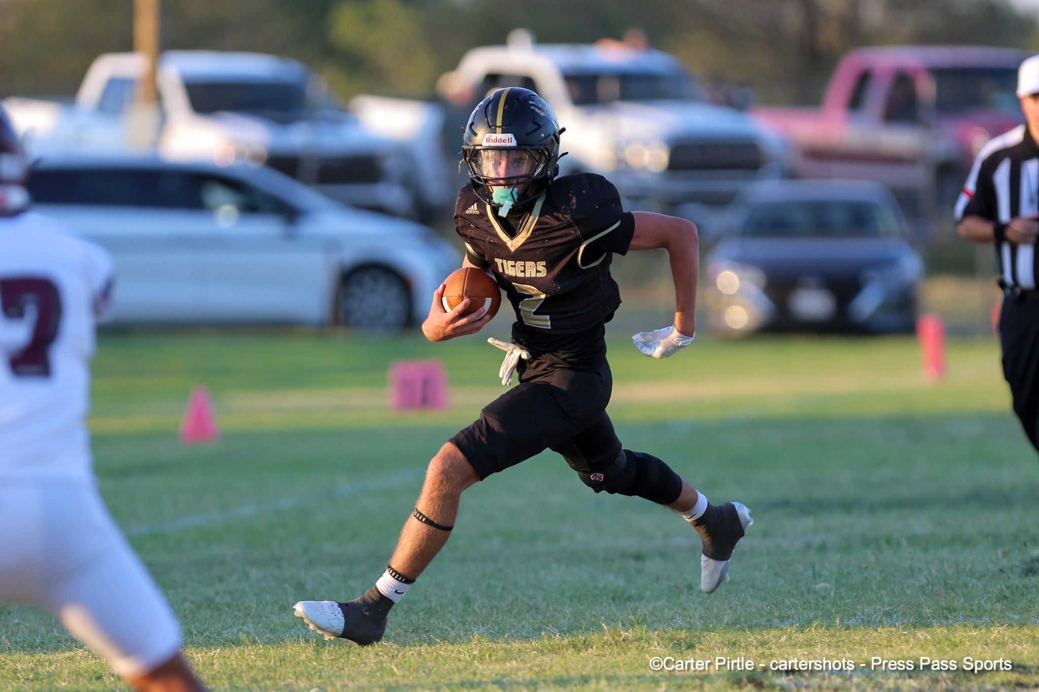 Football player in a black and gold Tigers jersey runs with the ball during a game, with a blurred crowd and cars in the background.